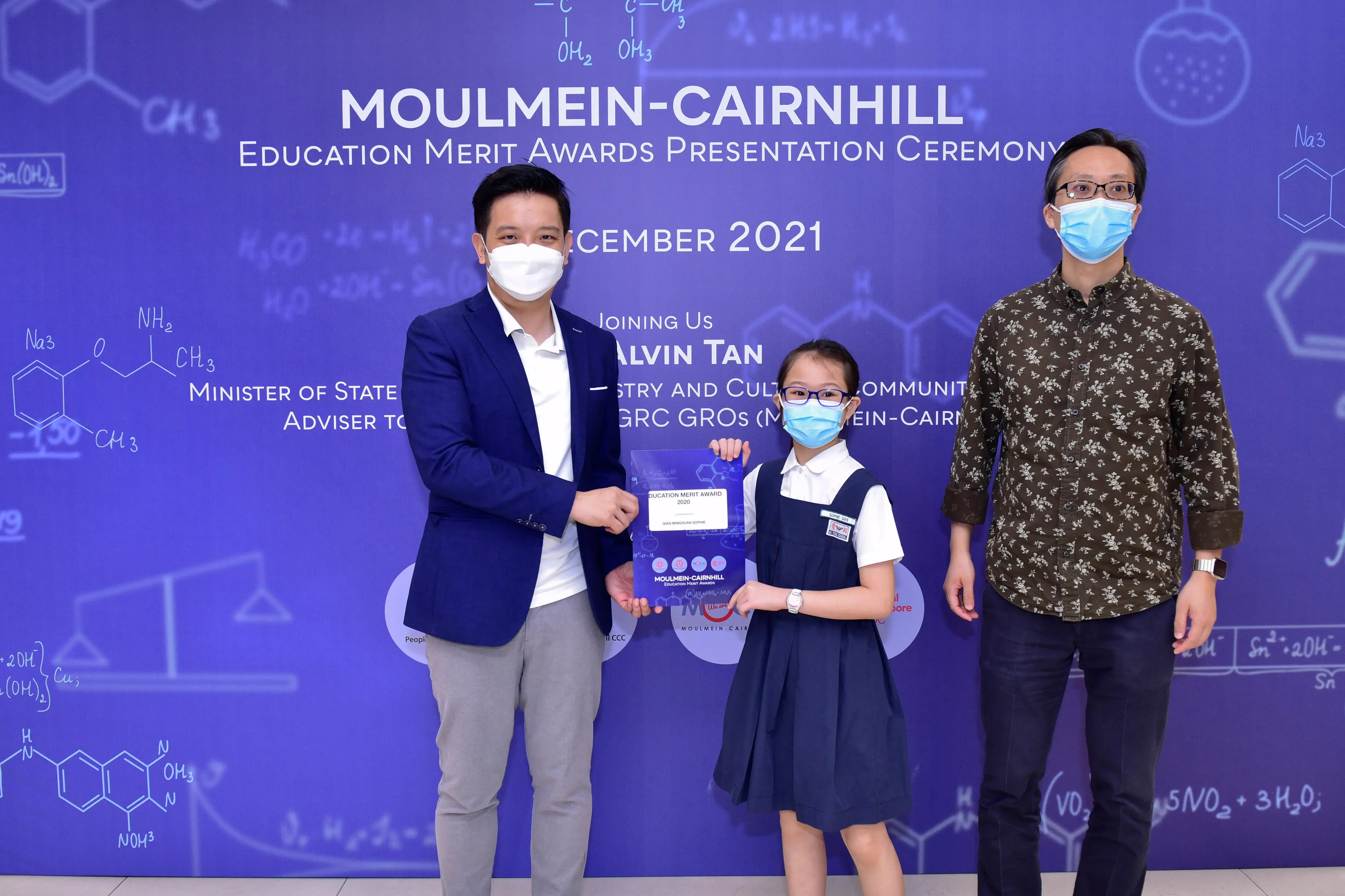 Two masked men and a young girl holding an award certificate at the Moulmein-Cairnhill Education Merit Awards 2021.