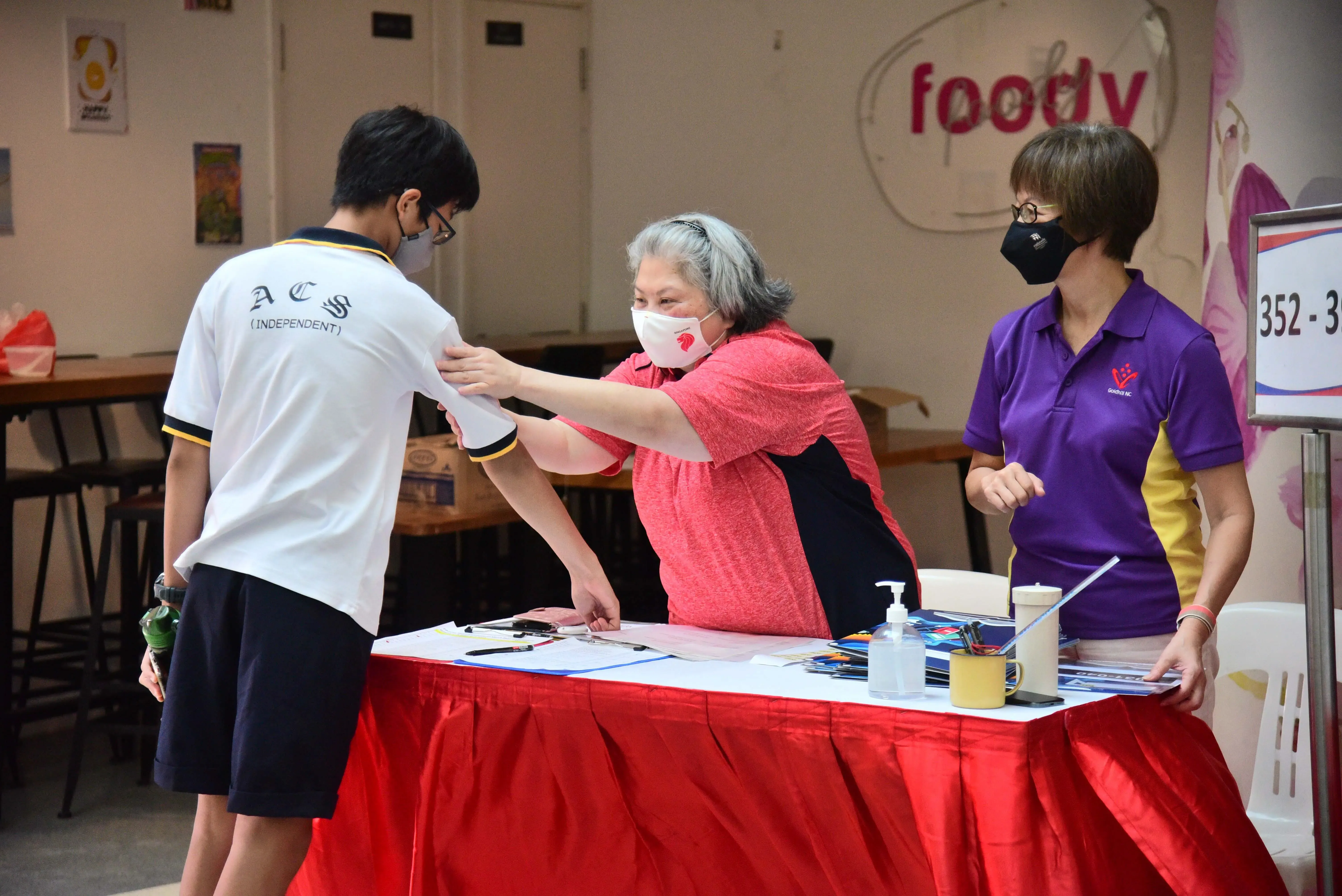 A masked student in an ACS uniform receives a document from a woman at a red-skirted registration table indoors.
