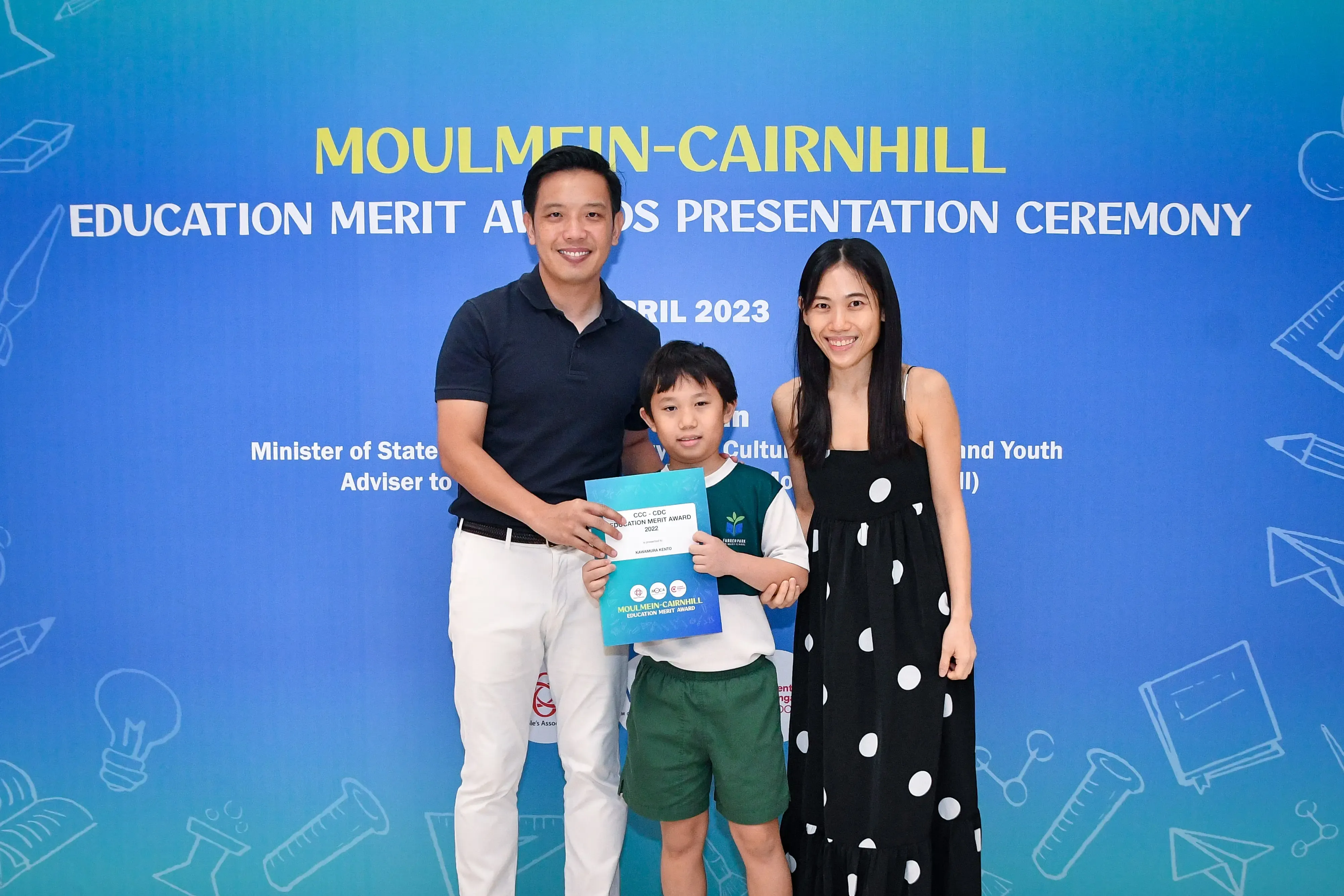 A boy holding an award certificate stands between two smiling adults at the Moulmein-Cairnhill Education Merit Awards 2023.
