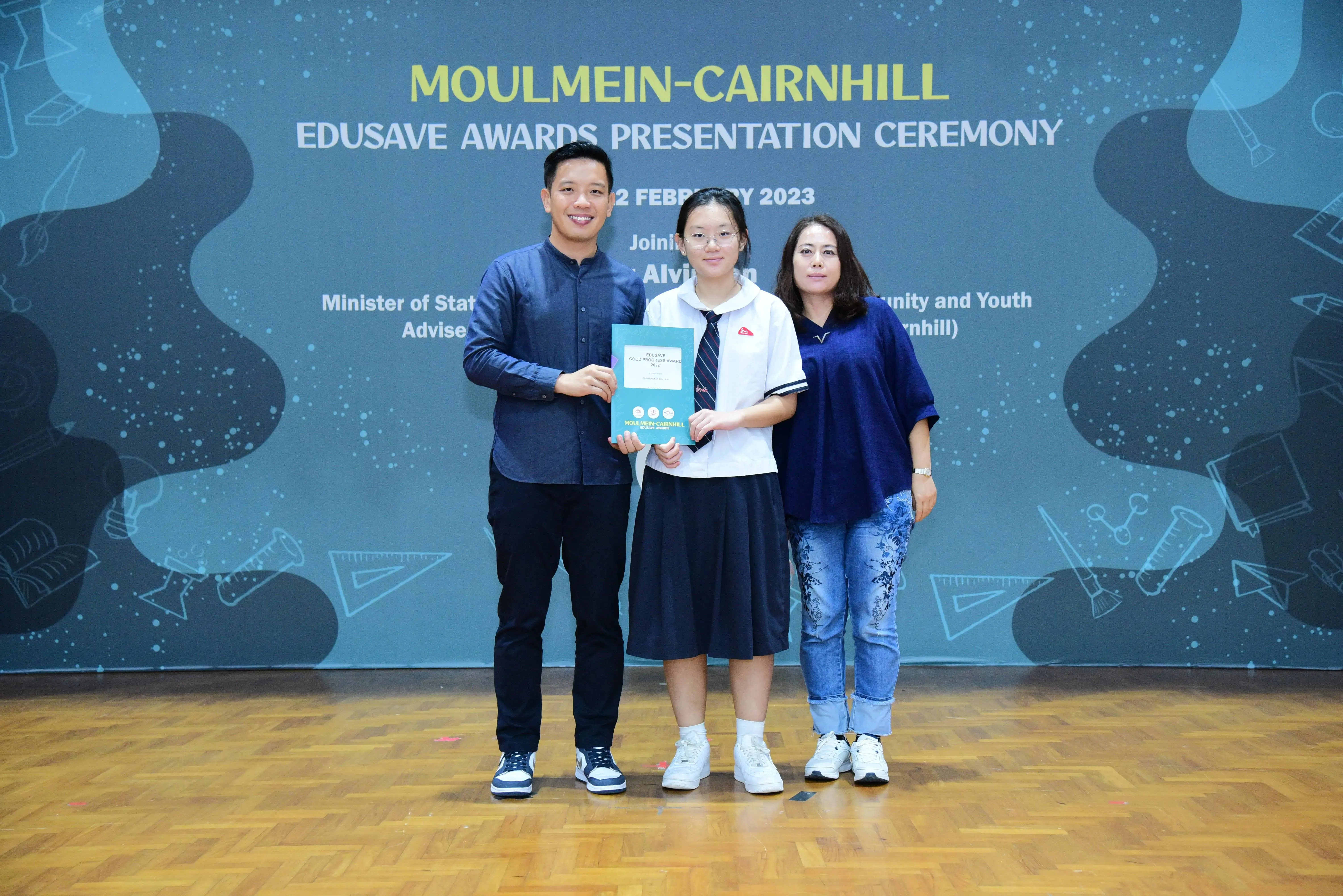 Three people pose at the Moulmein-Cairnhill Edusave Awards Presentation Ceremony, with a student holding an award folder.