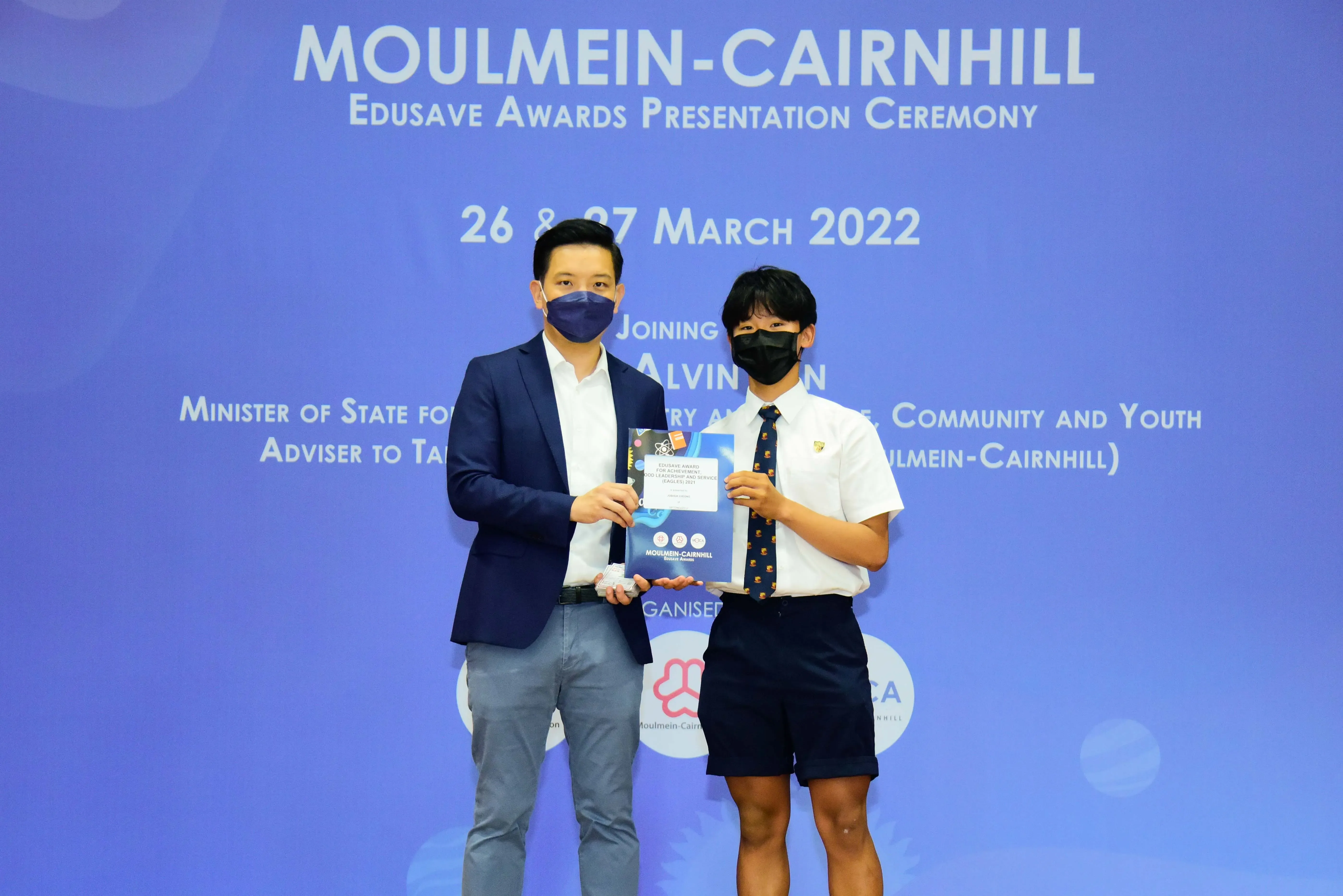Two masked individuals hold a certificate at the Moulmein-Cairnhill Edusave Awards Presentation Ceremony, March 2022.