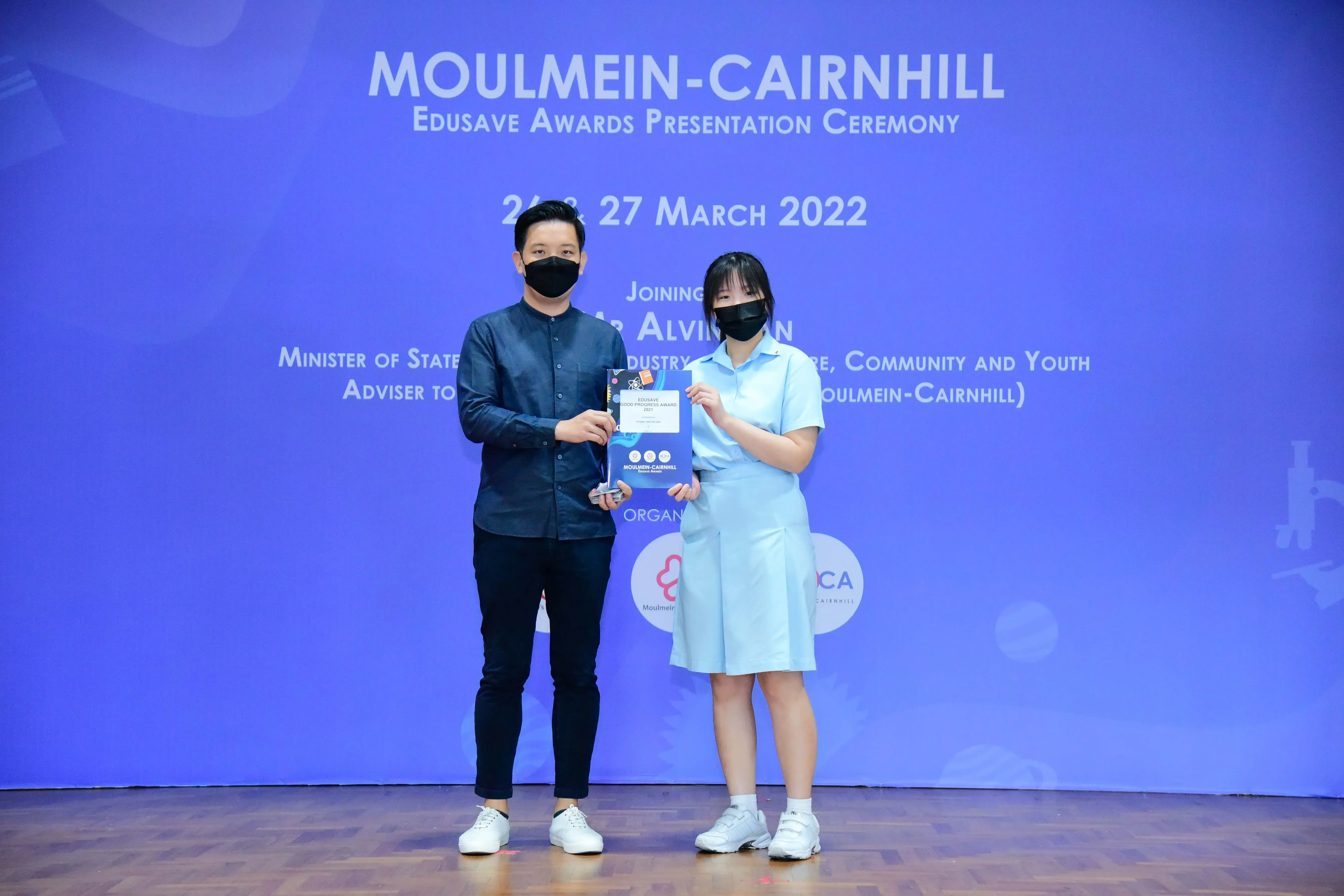Two masked people hold an award at the Moulmein-Cairnhill Edusave Awards Presentation Ceremony, March 2022.