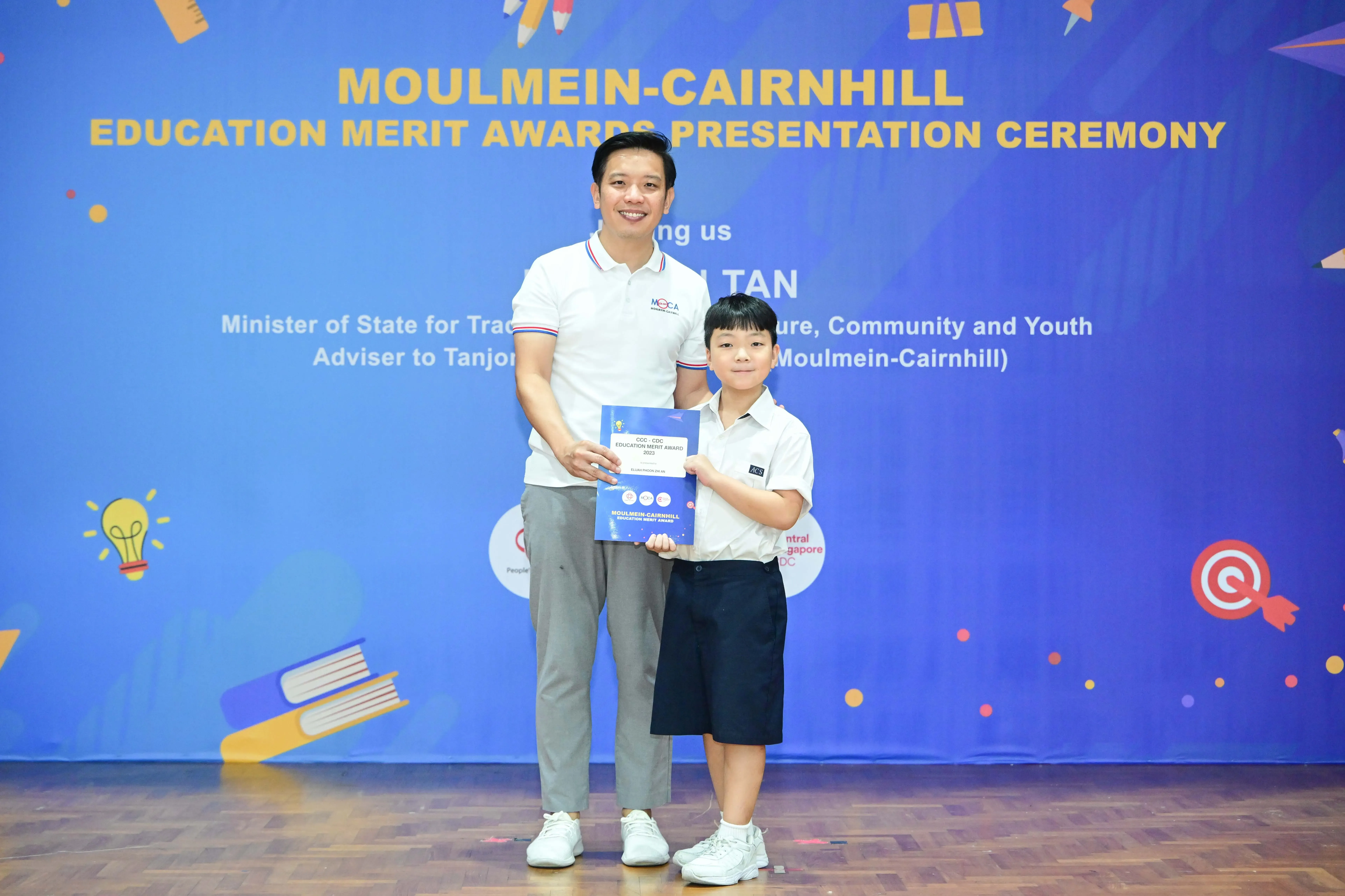 A man and a young boy hold a certificate in front of a Moulmein-Cairnhill Education Merit Awards banner.