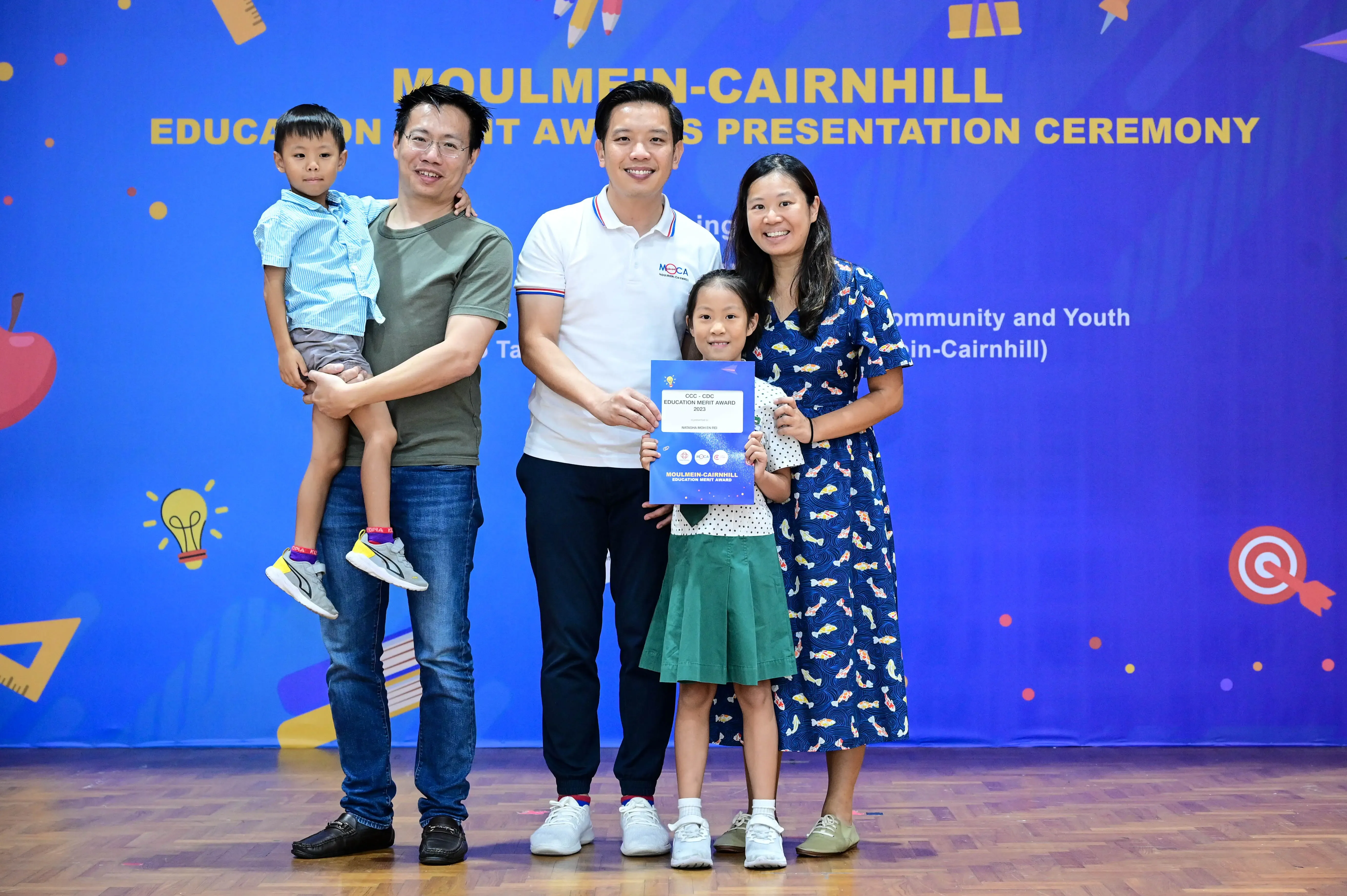 Four adults and two children pose at the Moulmein-Cairnhill Education Merit Awards Presentation Ceremony, girl holds certificate.