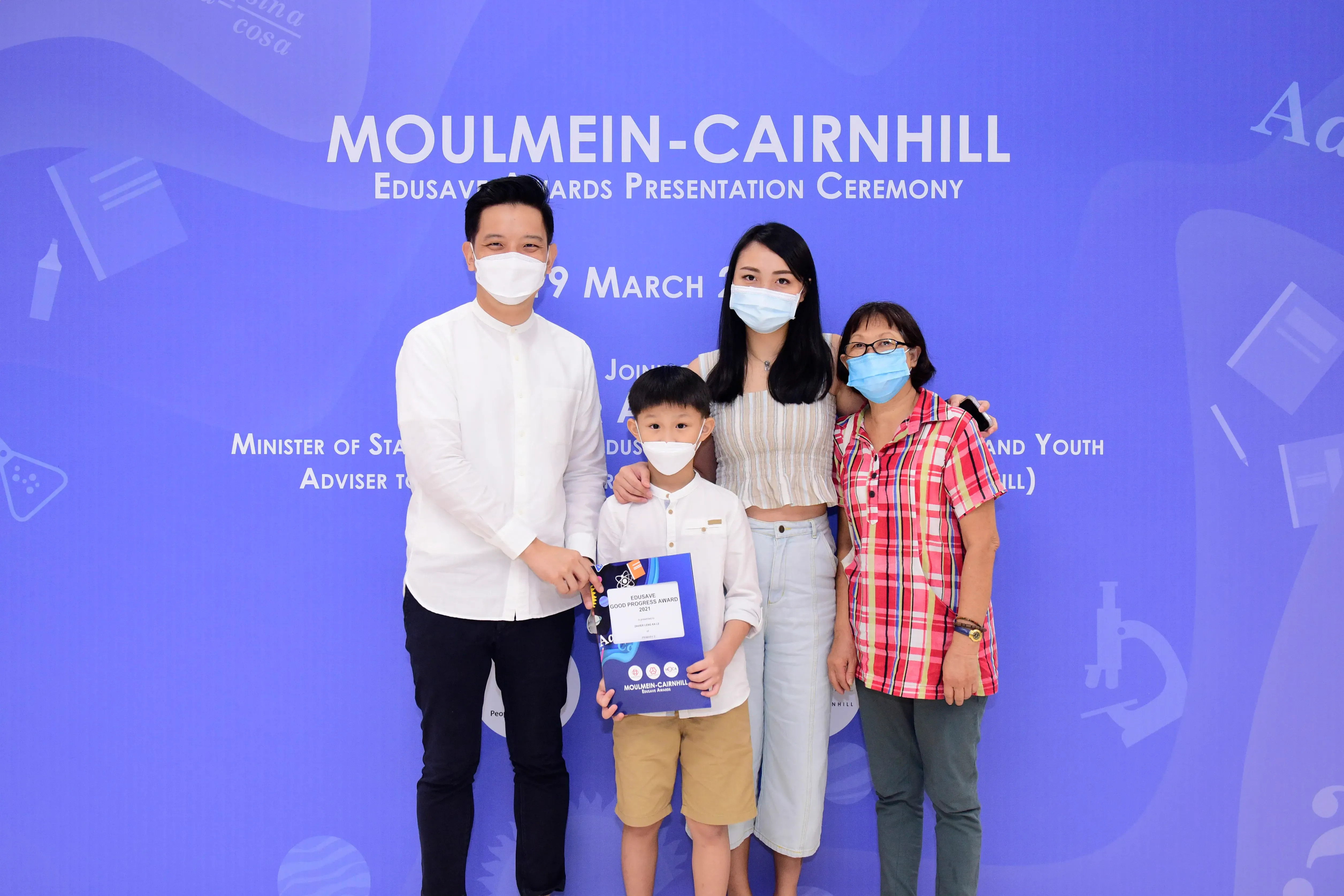 Four masked people pose at the Moulmein-Cairnhill Edusave Awards Ceremony, with a young boy holding an award folder.