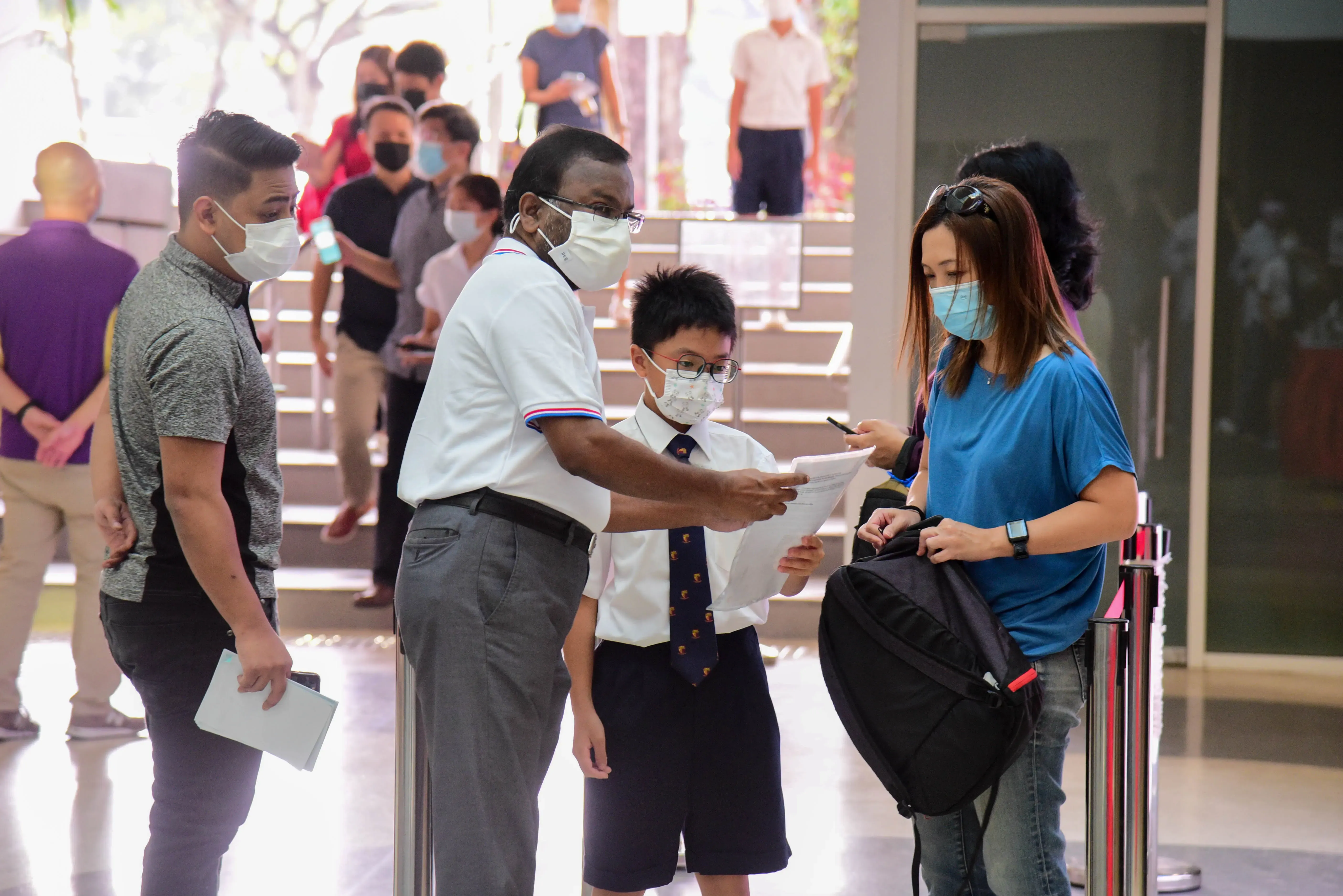 A masked man hands papers to a woman while a uniformed boy stands nearby, with a crowd of masked people in the background.