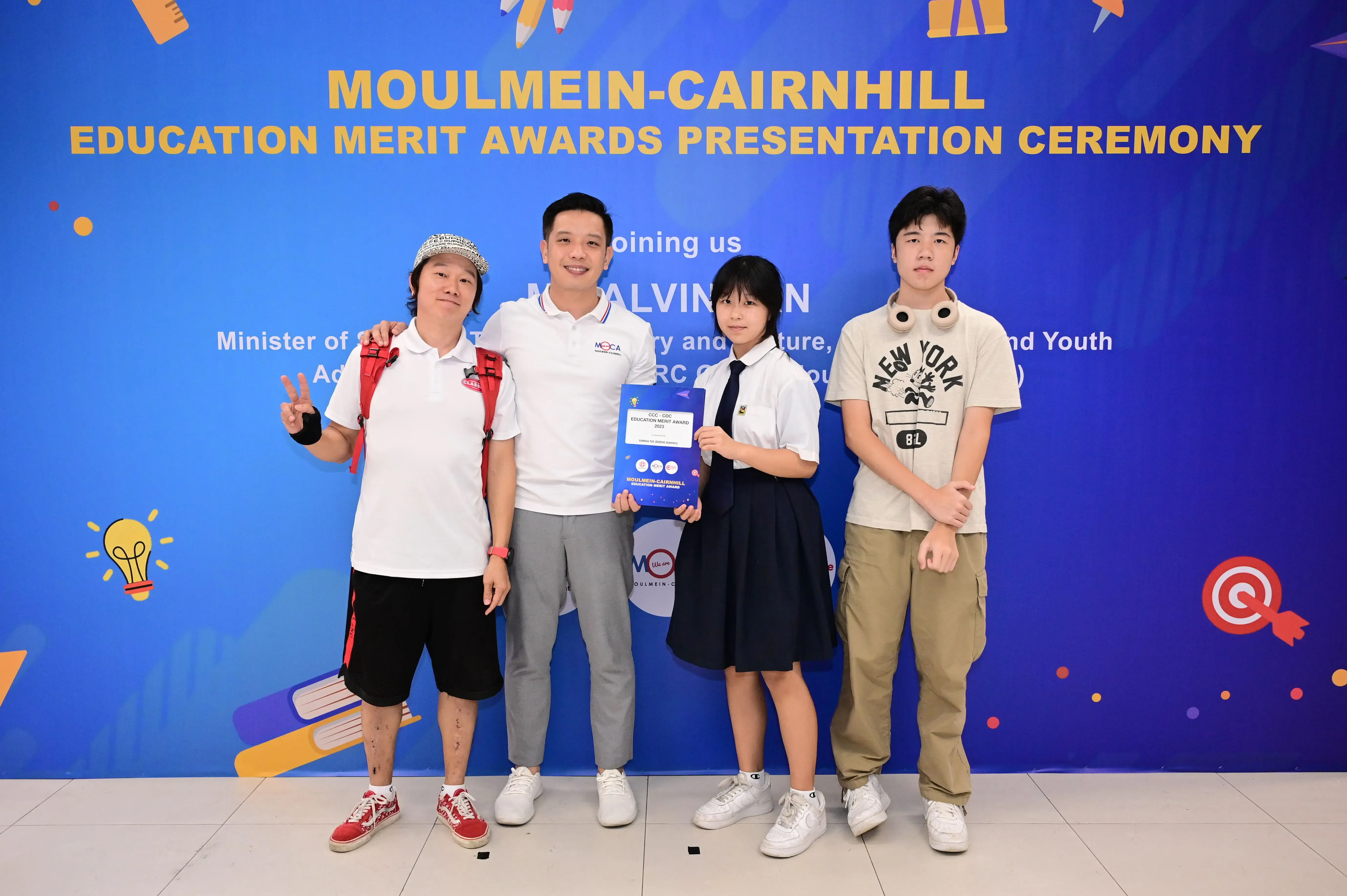 Four people posing in front of a Moulmein-Cairnhill Education Merit Awards Presentation Ceremony banner.
