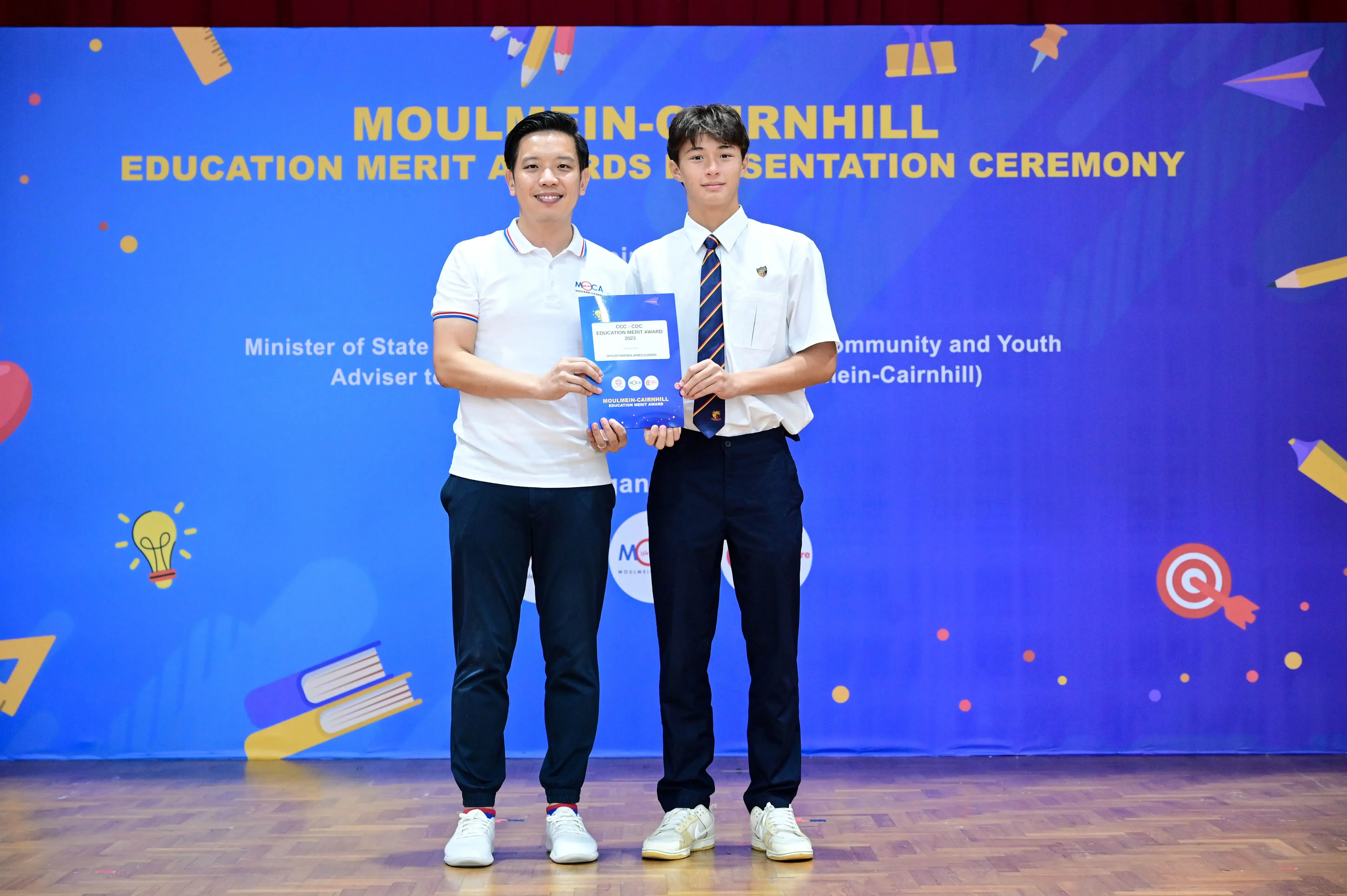 Two men hold an award certificate on stage at the Moulmein-Cairnhill Education Merit Awards Presentation Ceremony.