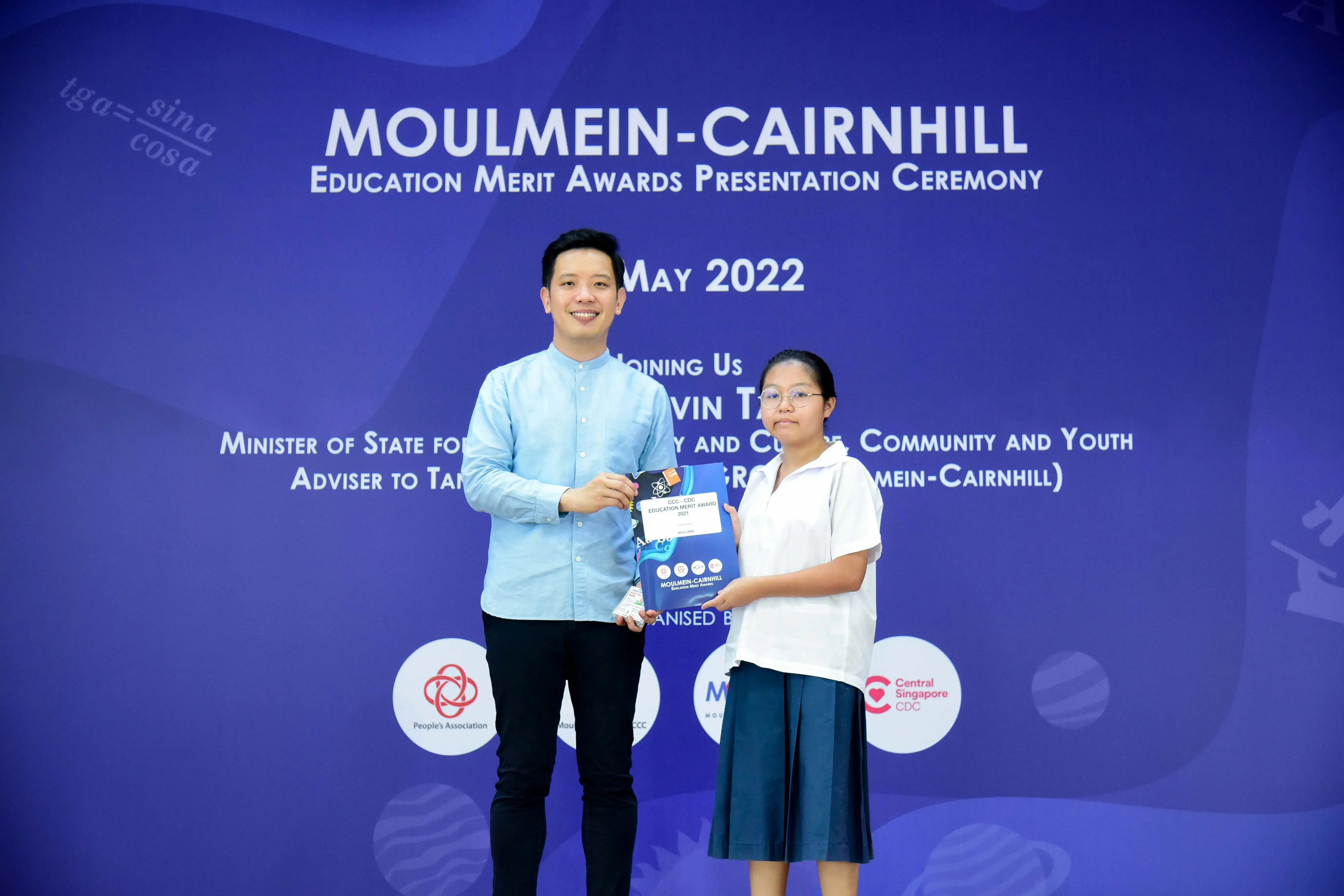 A man and a young girl hold an award together in front of a Moulmein-Cairnhill Education Merit Awards May 2022 banner.