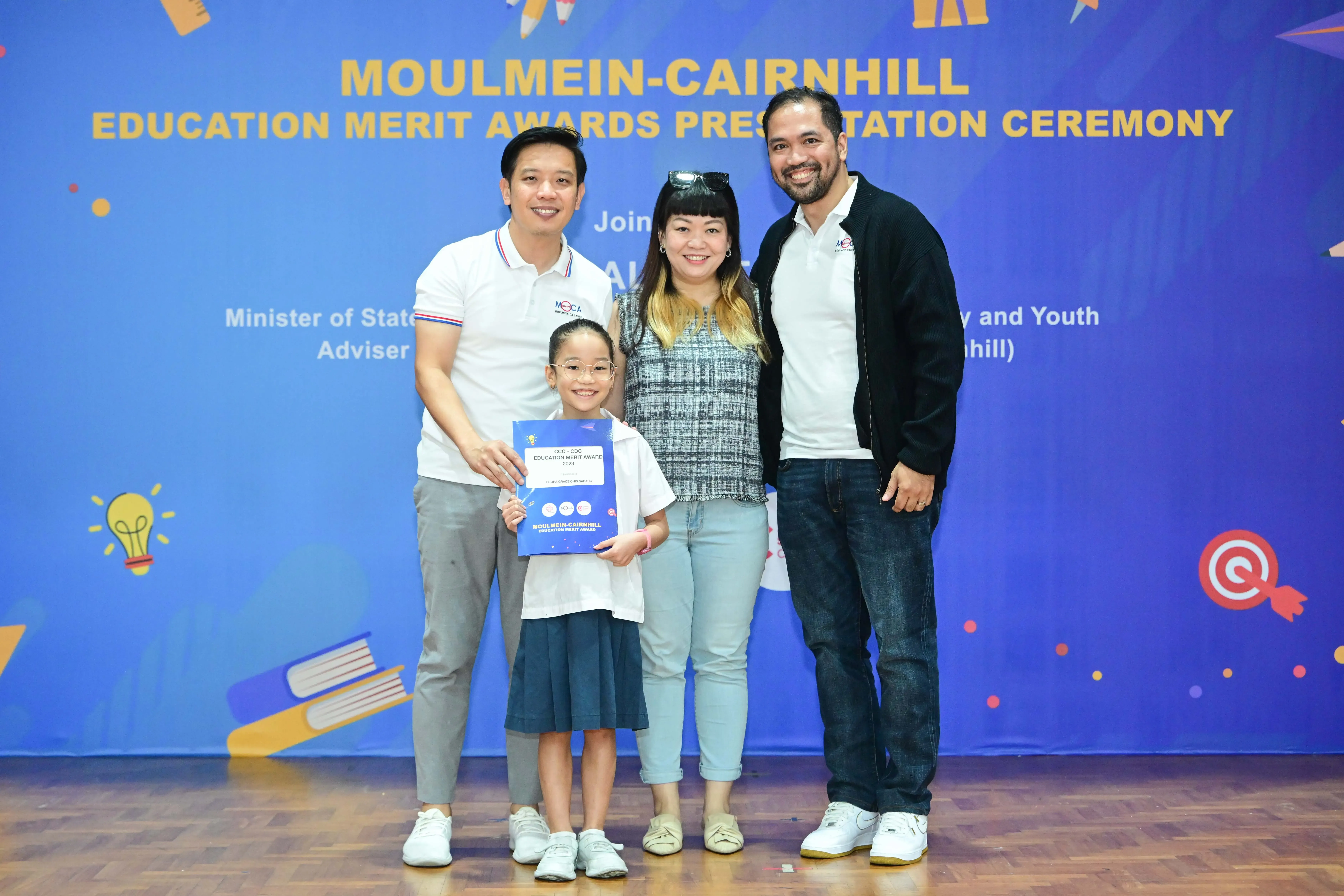 Three adults and a young girl holding an award certificate pose at the Moulmein-Cairnhill Education Merit Awards ceremony.