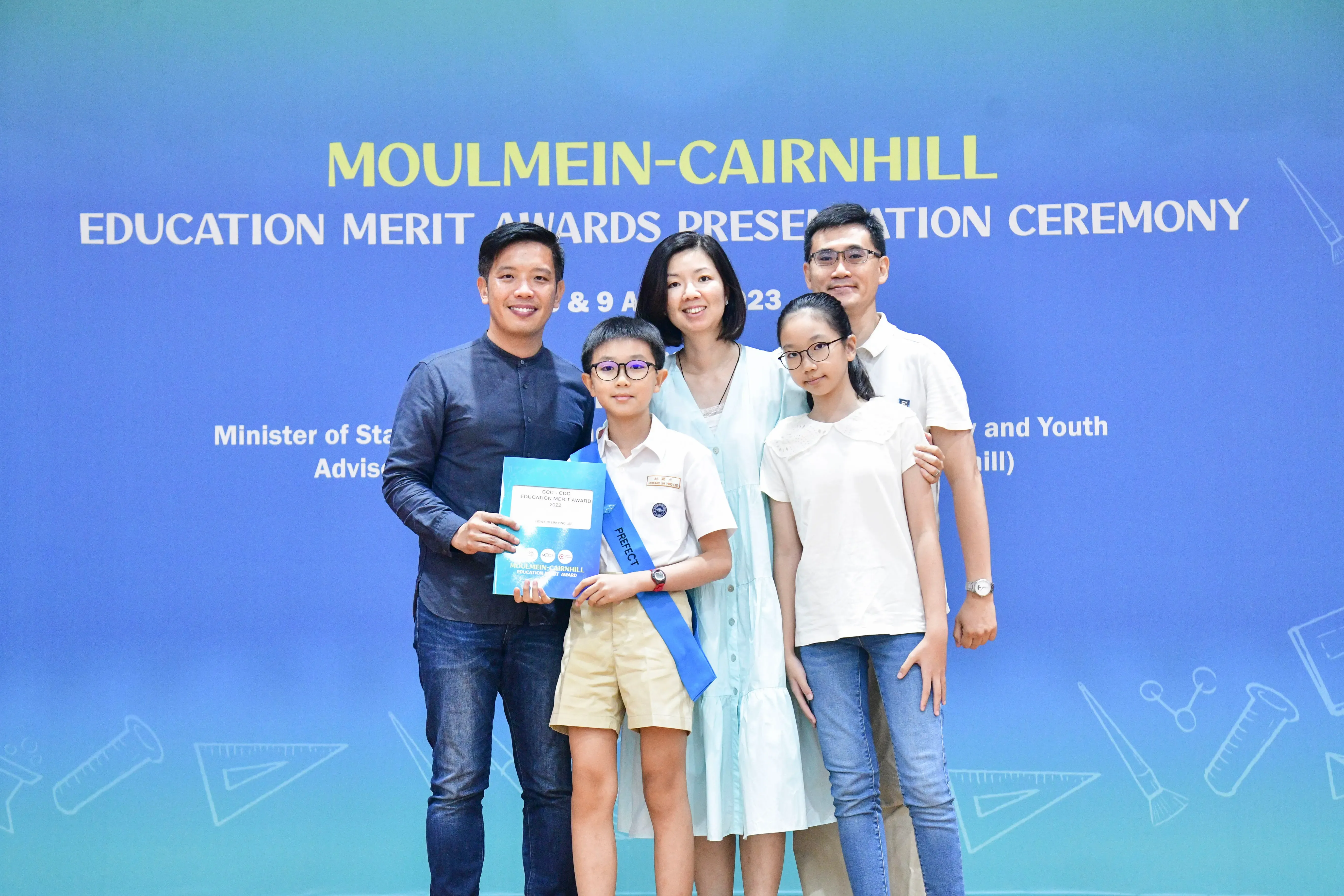Five people pose at the Moulmein-Cairnhill Education Merit Awards ceremony, including a boy with a blue sash holding an award.