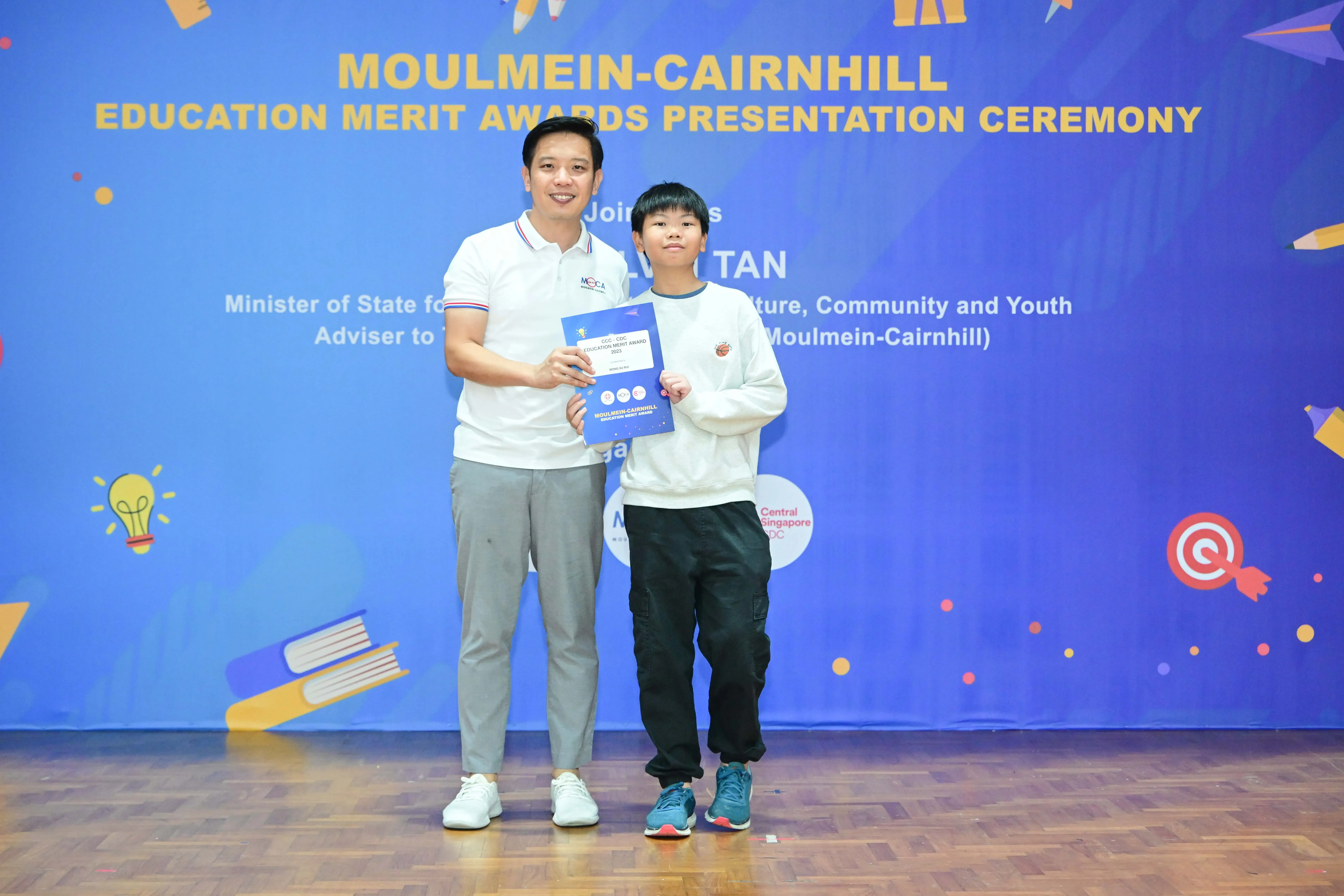 An adult and a child hold an award certificate in front of a Moulmein-Cairnhill Education Merit Awards banner.
