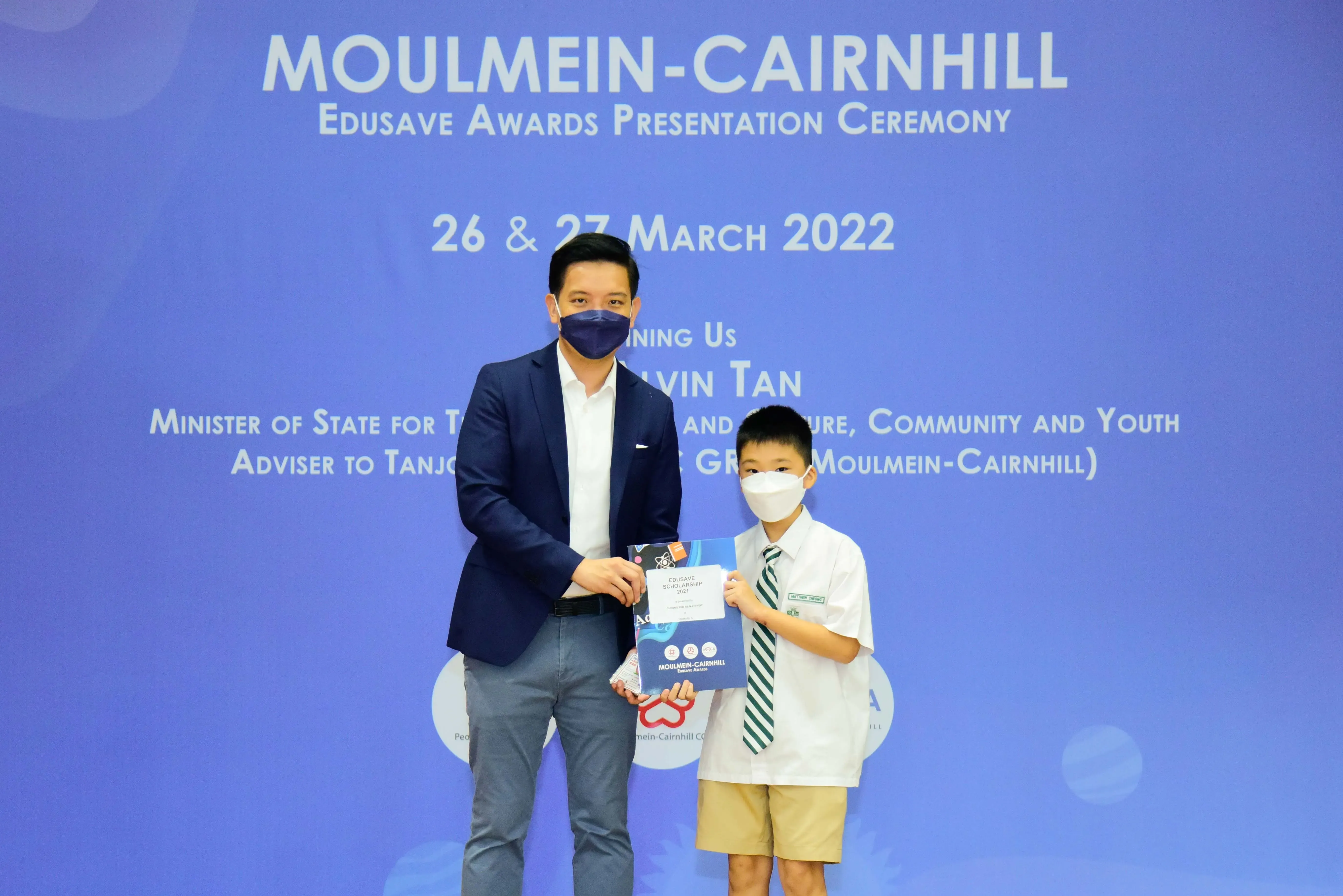 An adult and a young student in school uniform hold an Edusave award at the Moulmein-Cairnhill ceremony, March 2022.