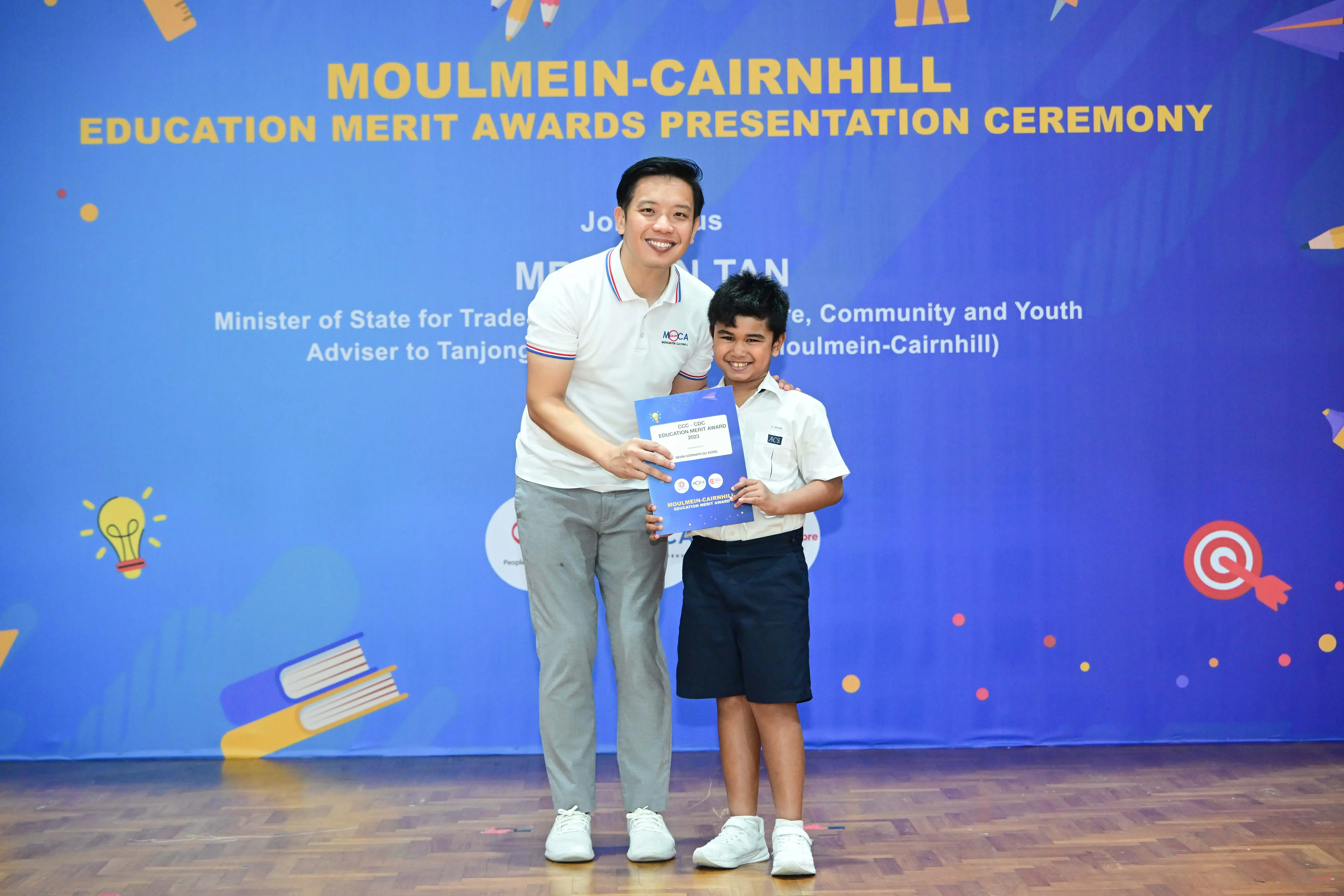 An adult and a young boy hold an award certificate at the Moulmein-Cairnhill Education Merit Awards Presentation Ceremony.