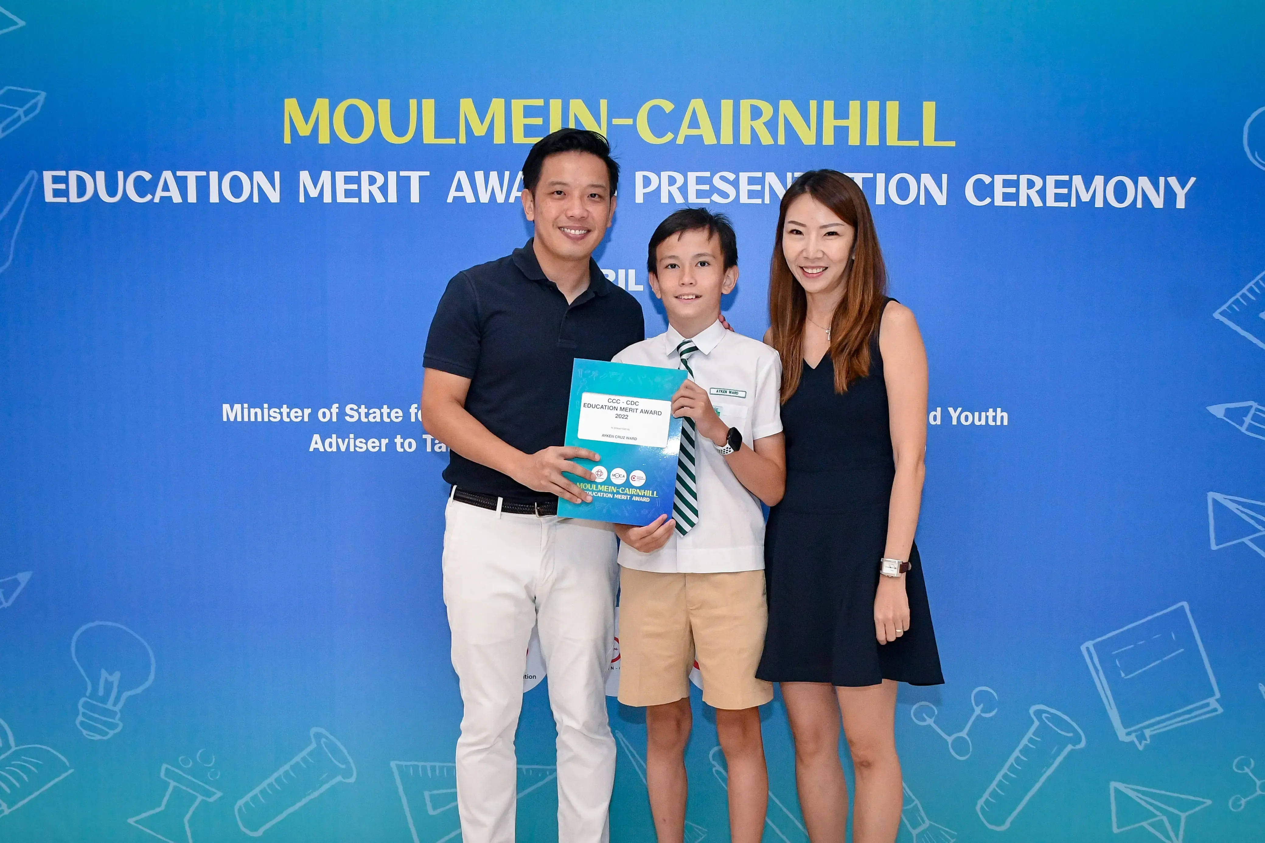 Two adults and a boy holding a certificate pose at the Moulmein-Cairnhill Education Merit Award Presentation Ceremony.