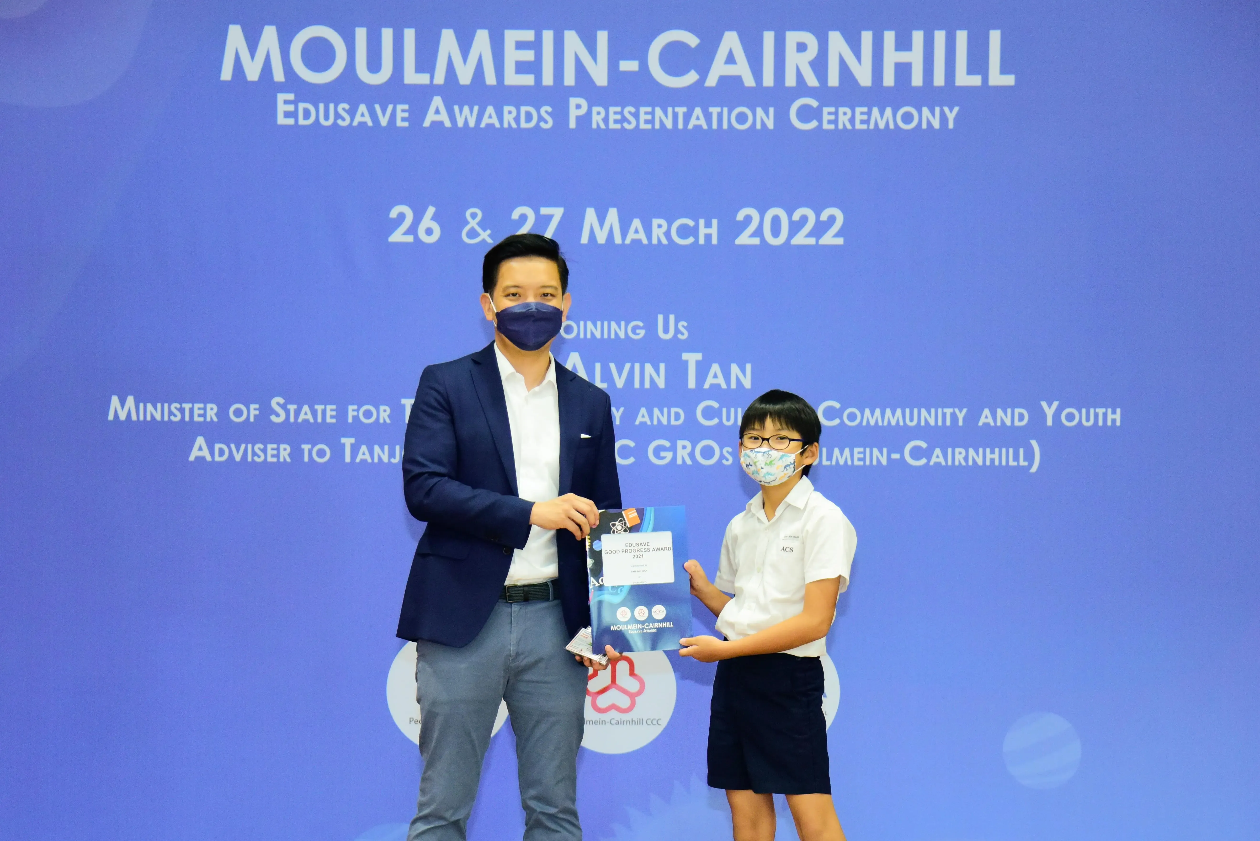 An adult in a blazer presents an award certificate to a young student at the Moulmein-Cairnhill Edusave Awards Ceremony 2022.
