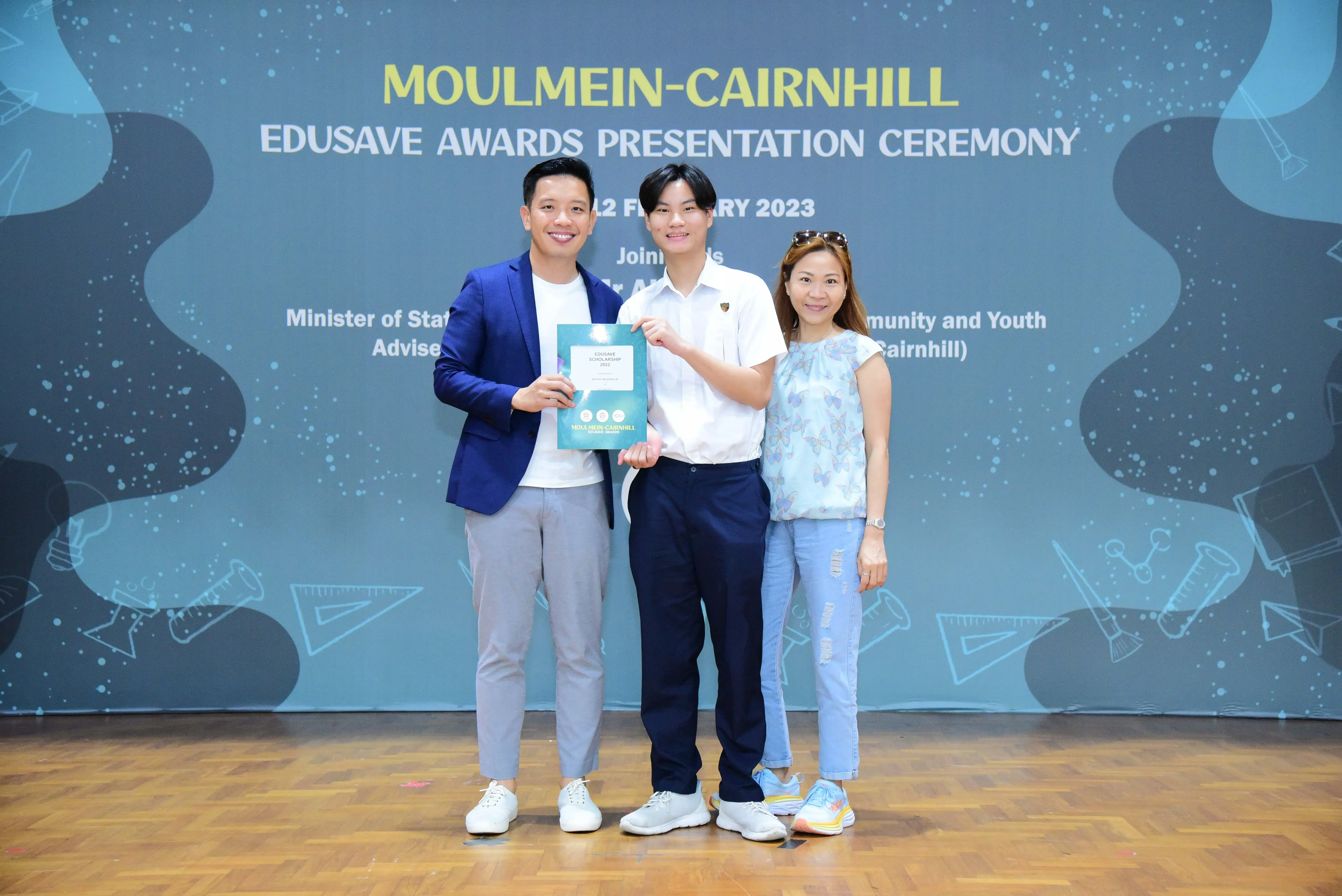 Three people stand smiling before a Moulmein-Cairnhill Edusave Awards Presentation Ceremony banner, holding a certificate.