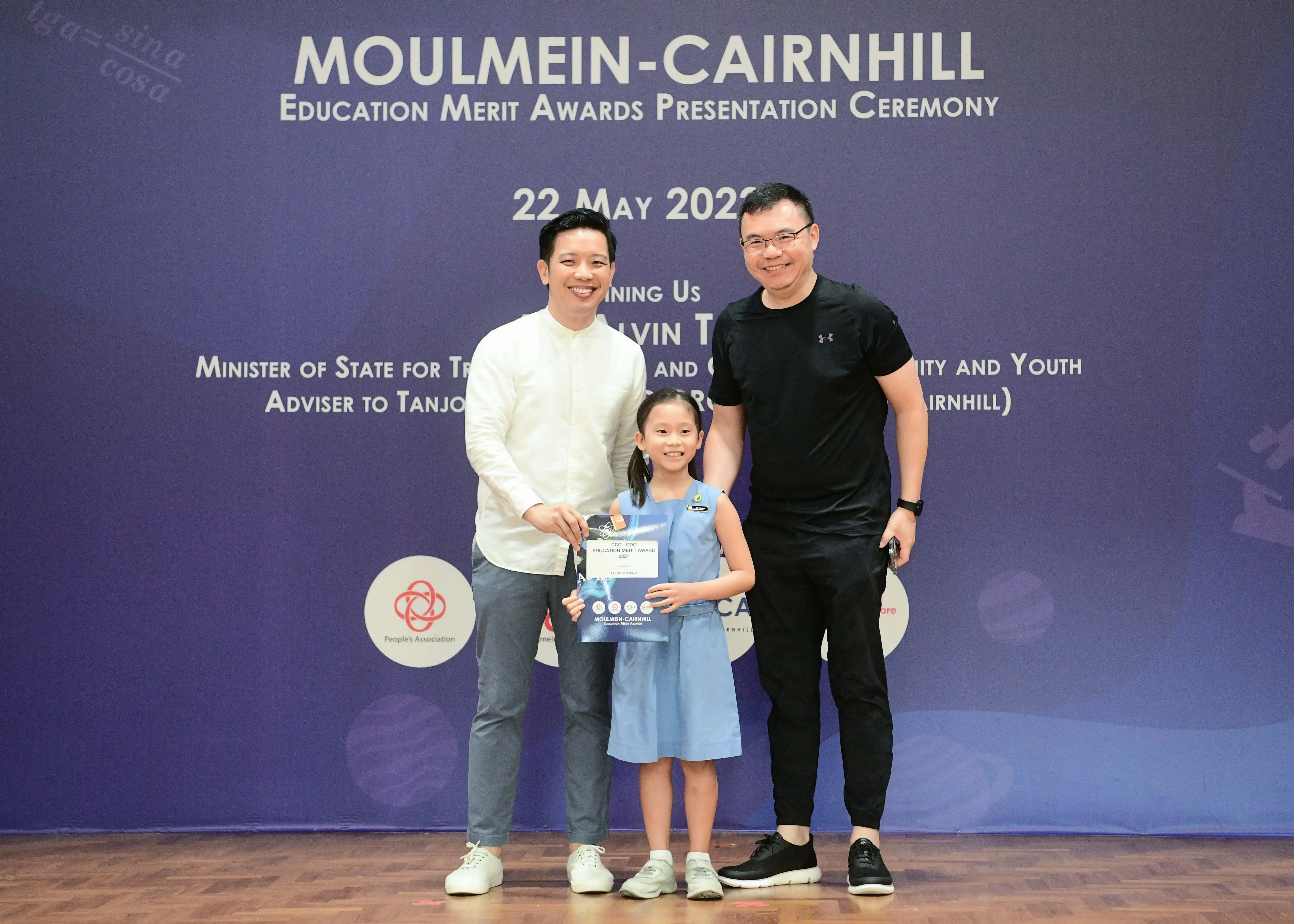Two men and a young girl holding an award certificate pose at the Moulmein-Cairnhill Education Merit Awards ceremony.