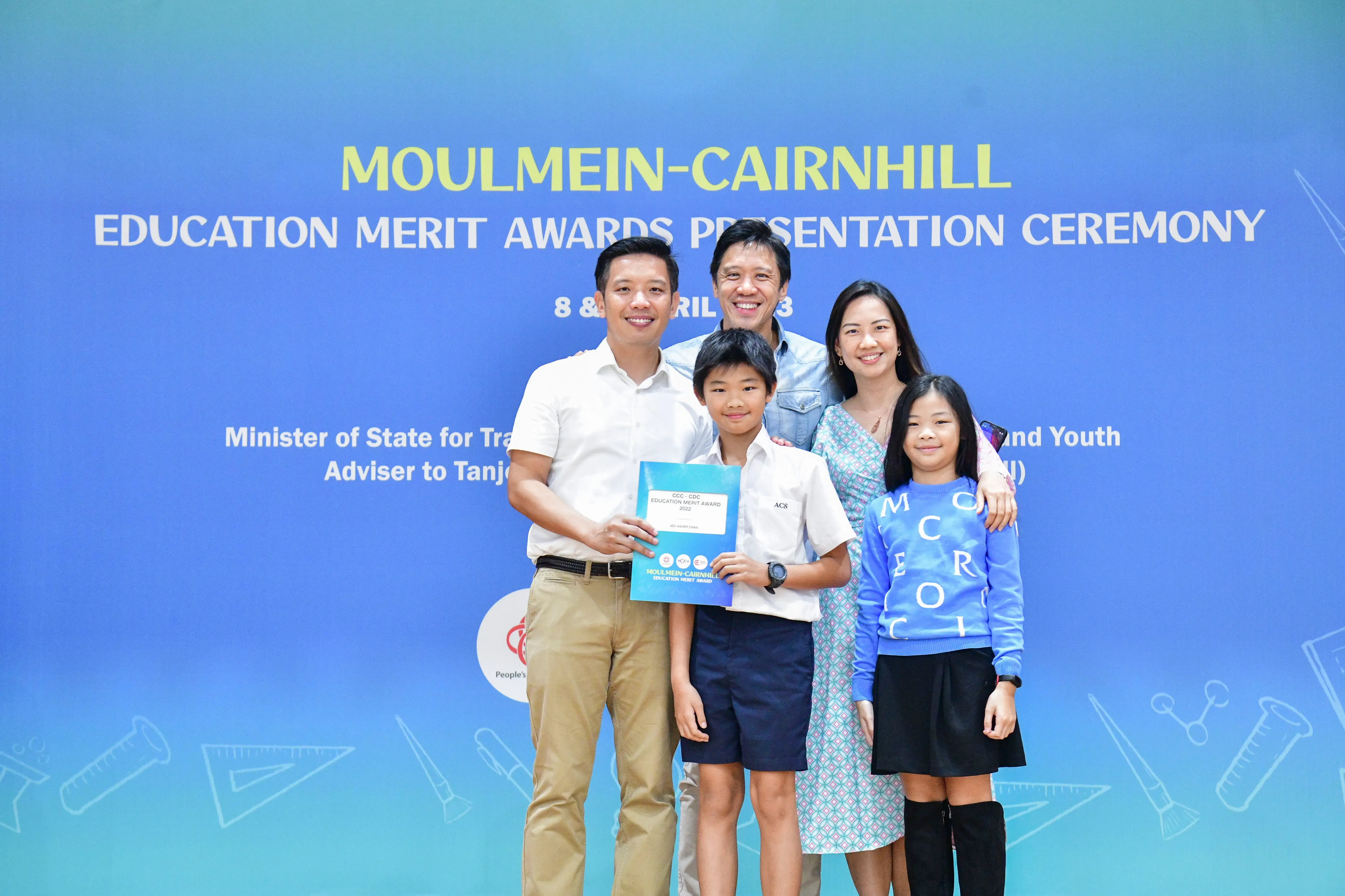 Five people pose smiling before a blue Moulmein-Cairnhill Education Merit Awards Presentation Ceremony banner, boy holds award.