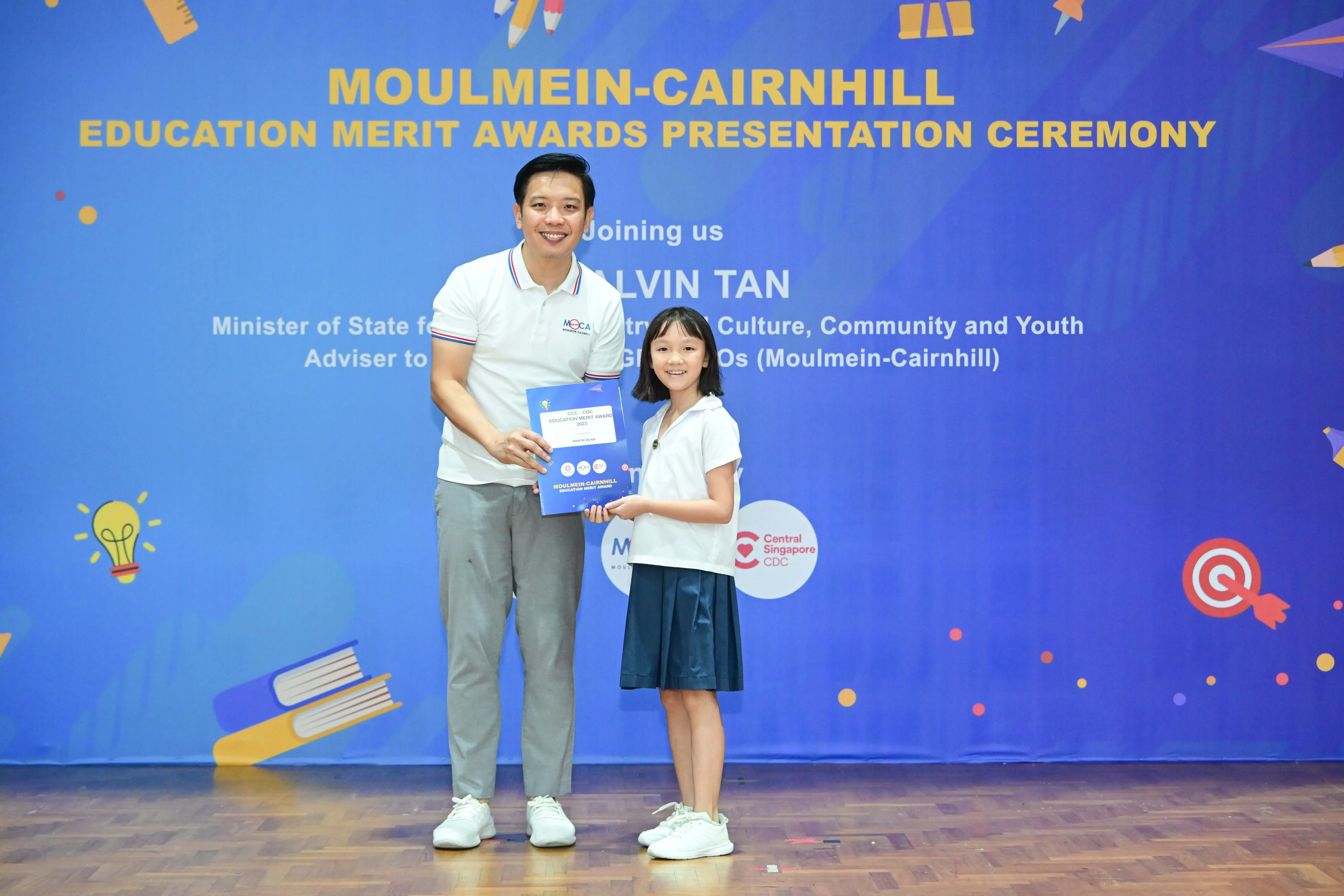 A man and a young girl hold a blue certificate together in front of a Moulmein-Cairnhill Education Merit Awards banner.