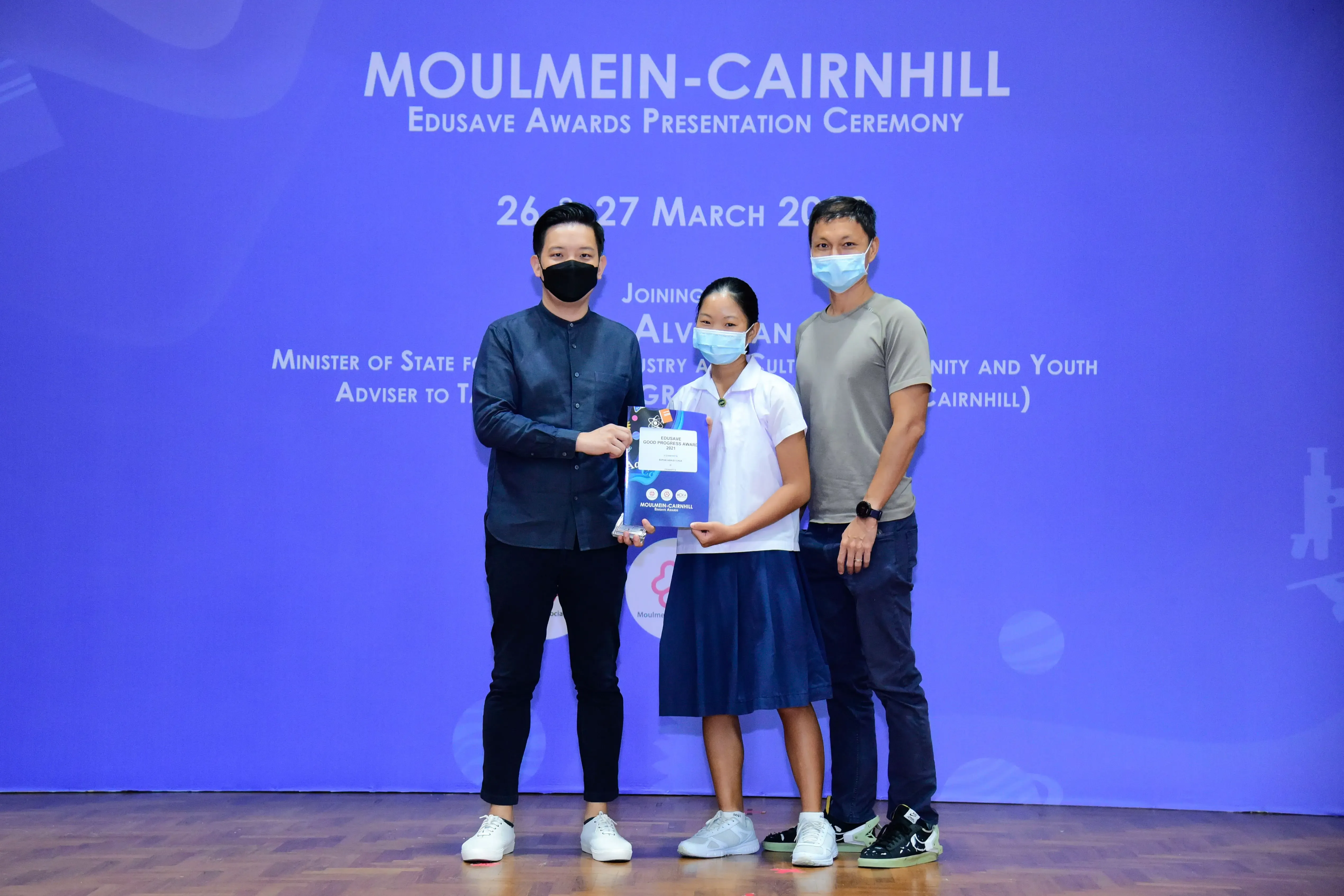 Three masked people stand before a blue Moulmein-Cairnhill Edusave Awards Presentation Ceremony banner, holding a certificate.