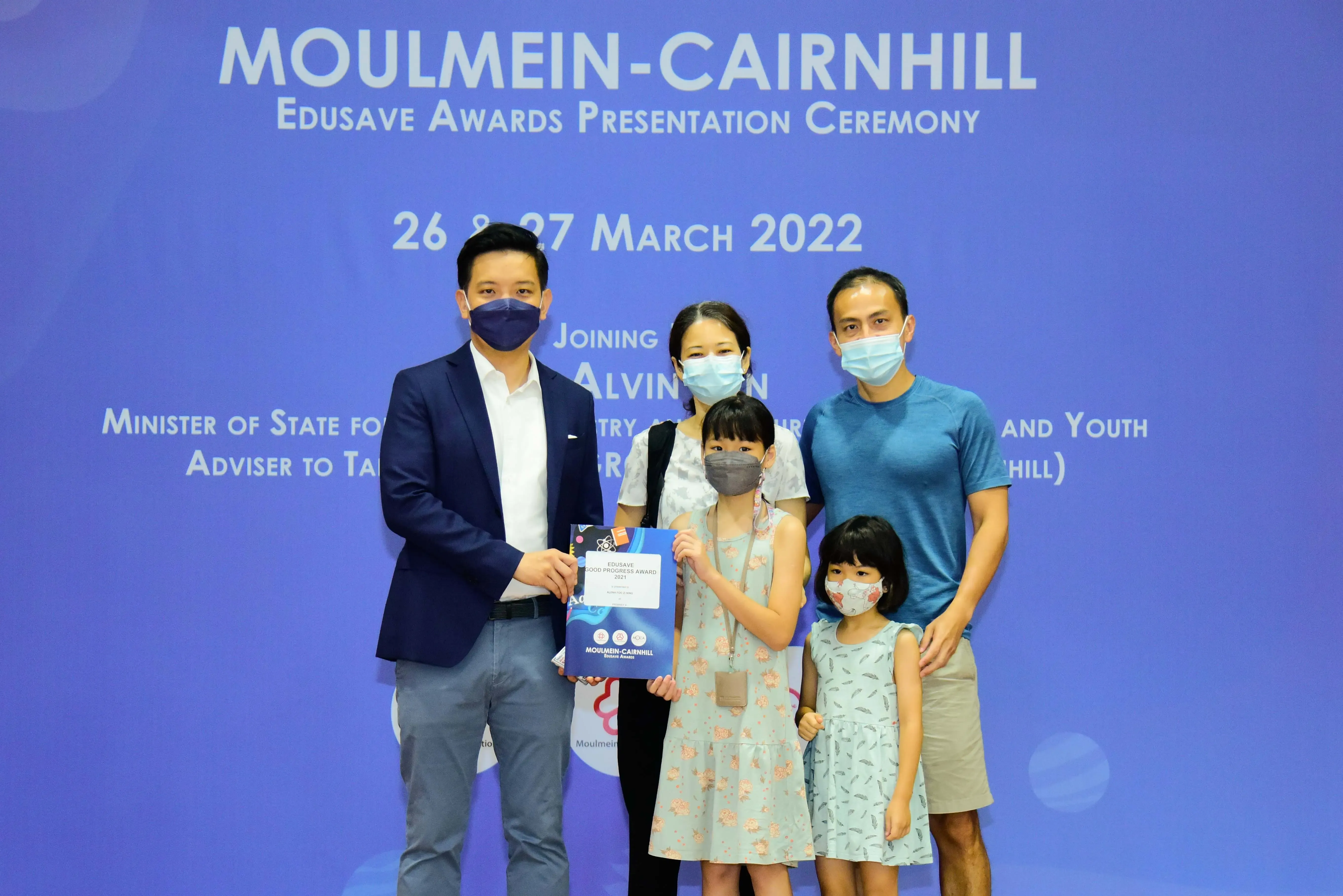 Five masked people pose before a Moulmein-Cairnhill Edusave Awards Presentation Ceremony banner dated March 2022.