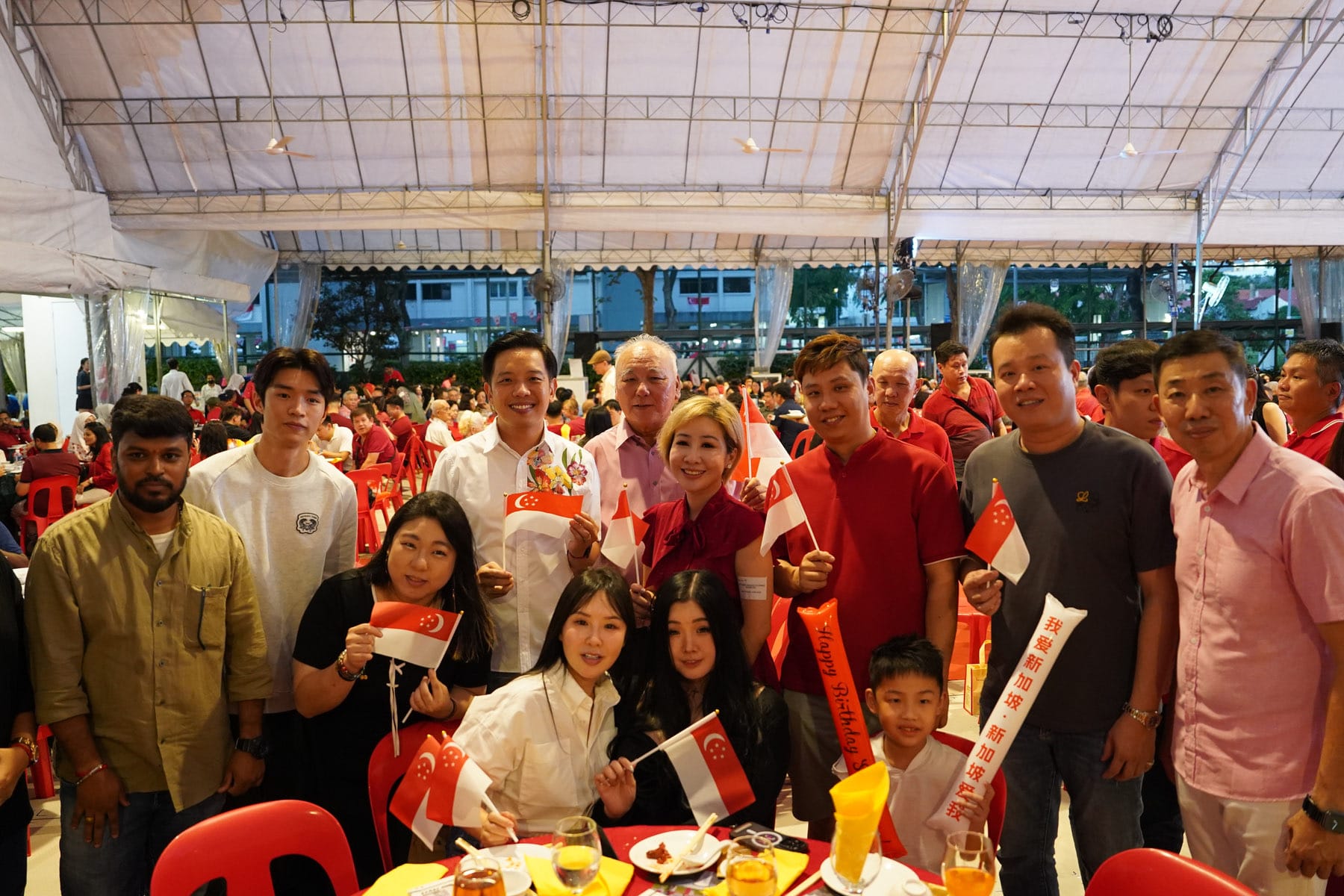 A group of smiling people holding small Singapore flags at an outdoor celebration under a white tent.