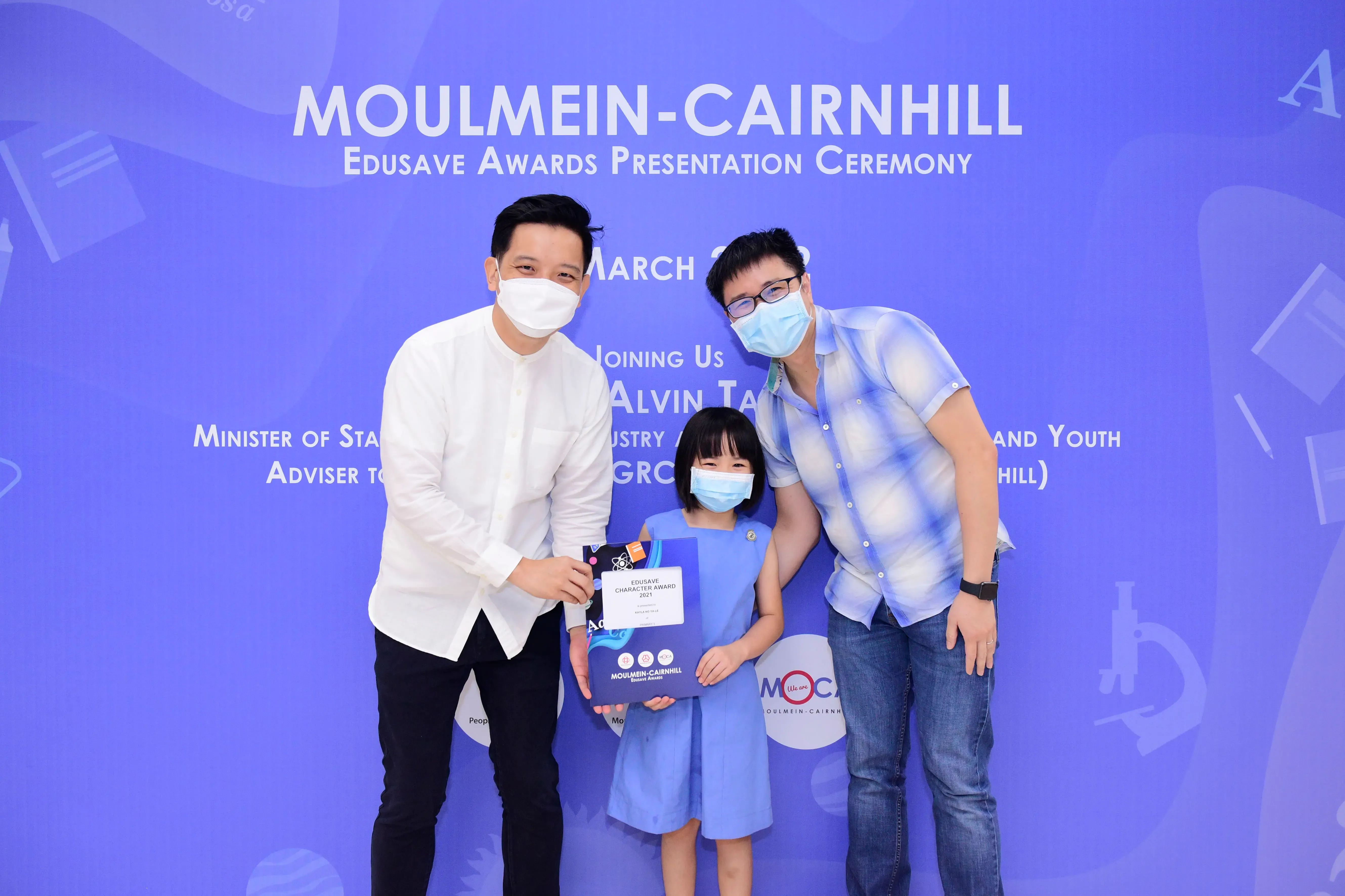 Two masked men and a masked girl holding an award certificate at the Moulmein-Cairnhill Edusave Awards Ceremony.