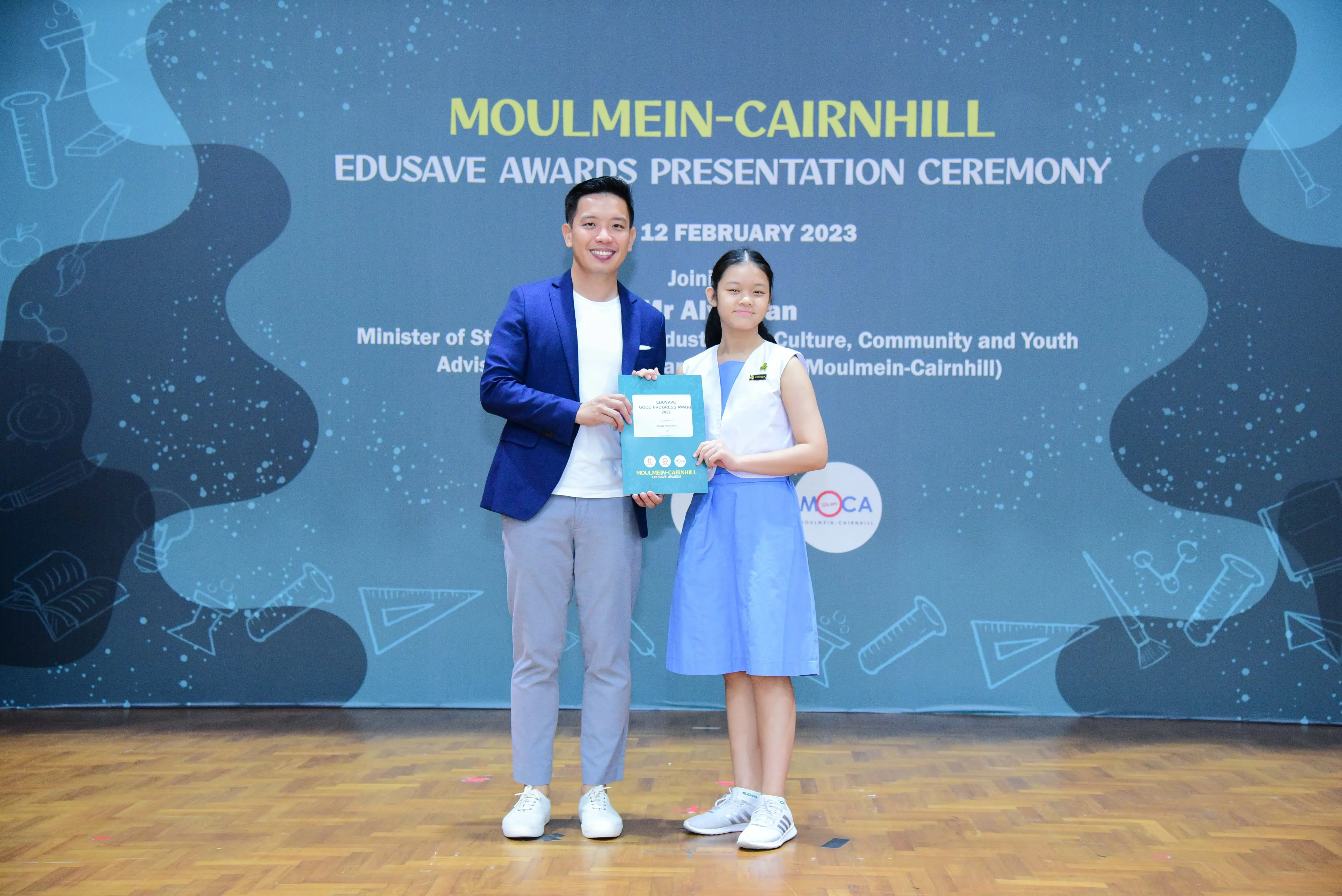 A man in a blue blazer and a young girl in a blue dress hold a certificate at the Moulmein-Cairnhill Edusave Awards 2023.