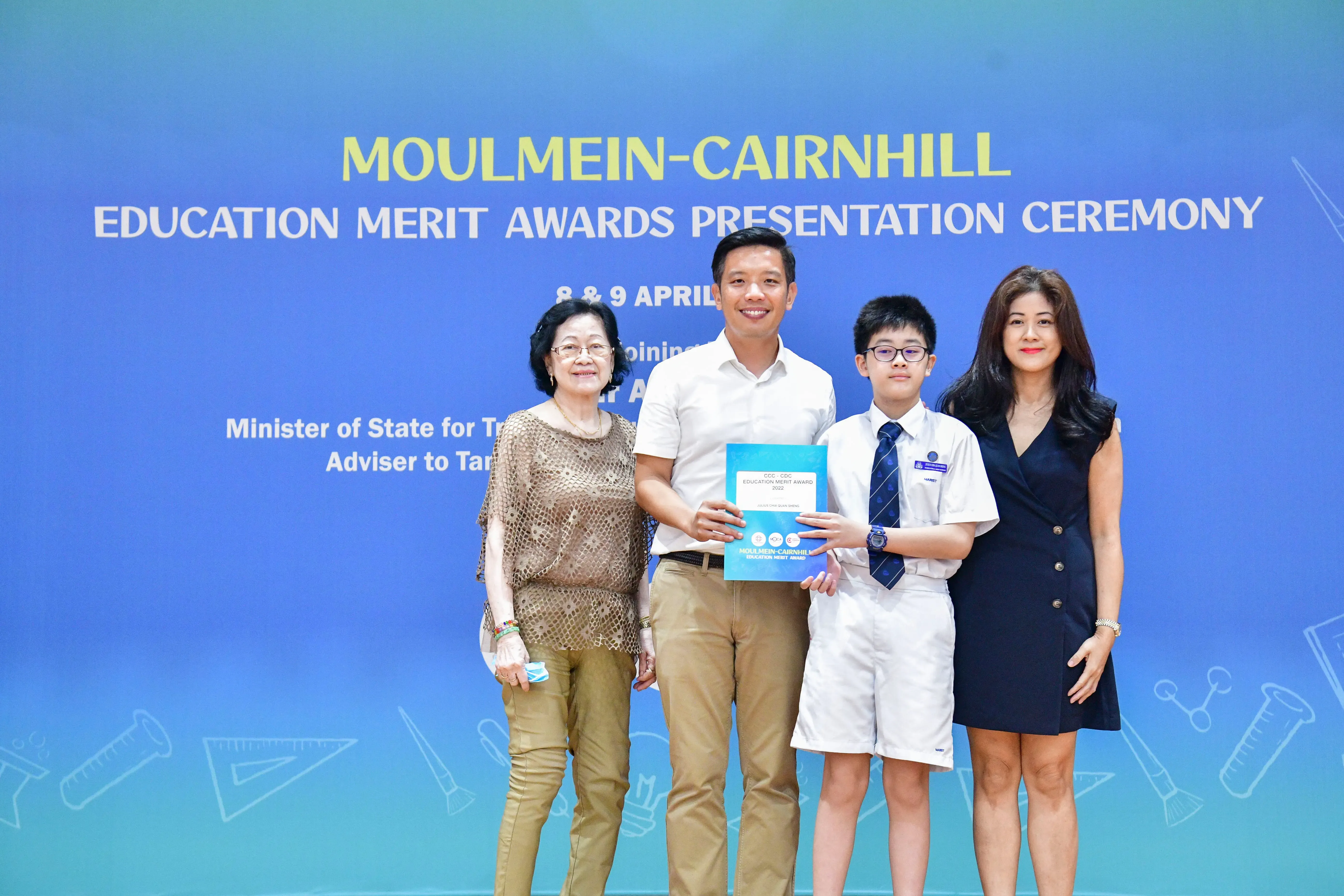 Four people pose at the Moulmein-Cairnhill Education Merit Awards Presentation Ceremony, with a boy holding an award certificate.