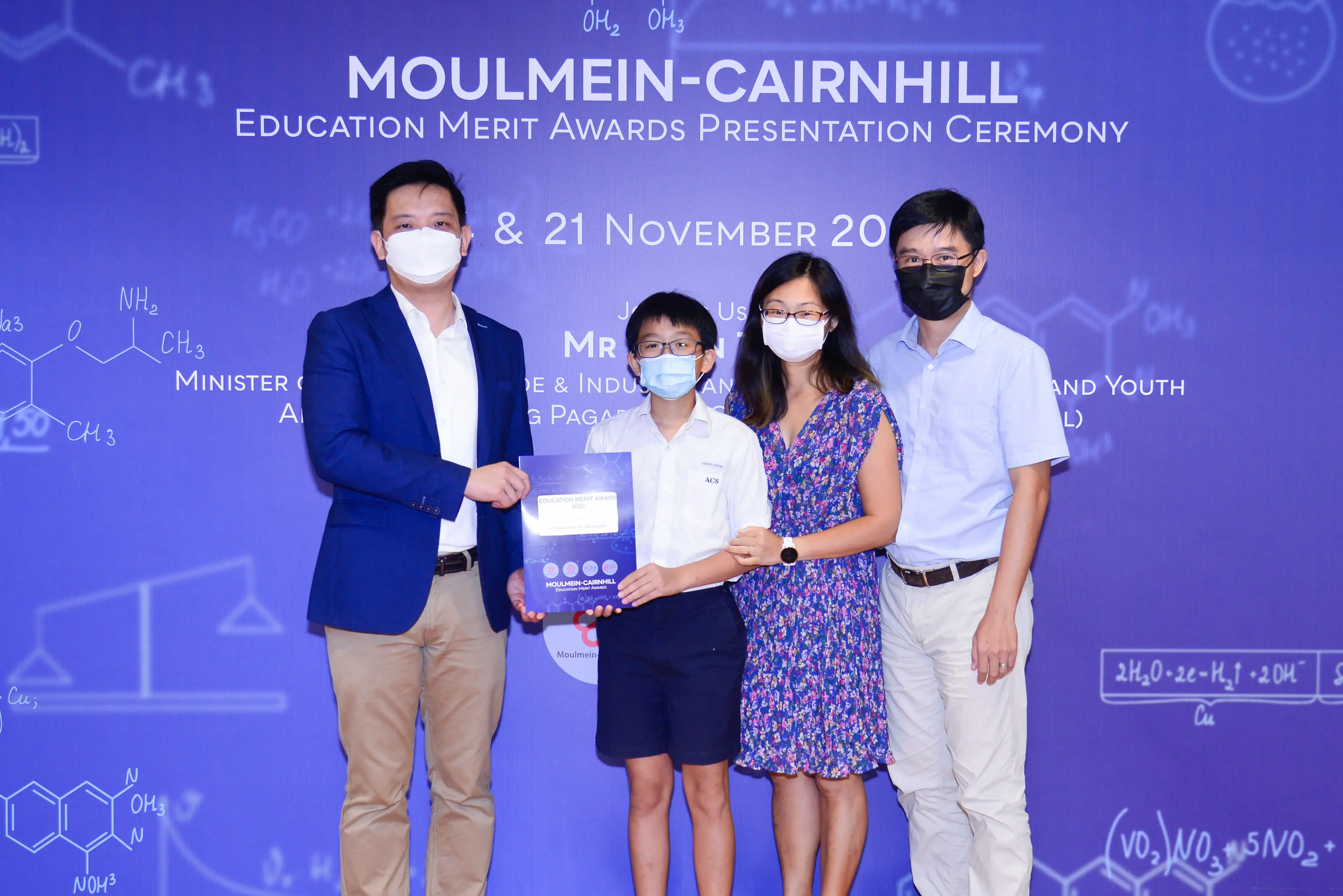 Four masked people pose at the Moulmein-Cairnhill Education Merit Awards ceremony, a boy holding an award certificate.
