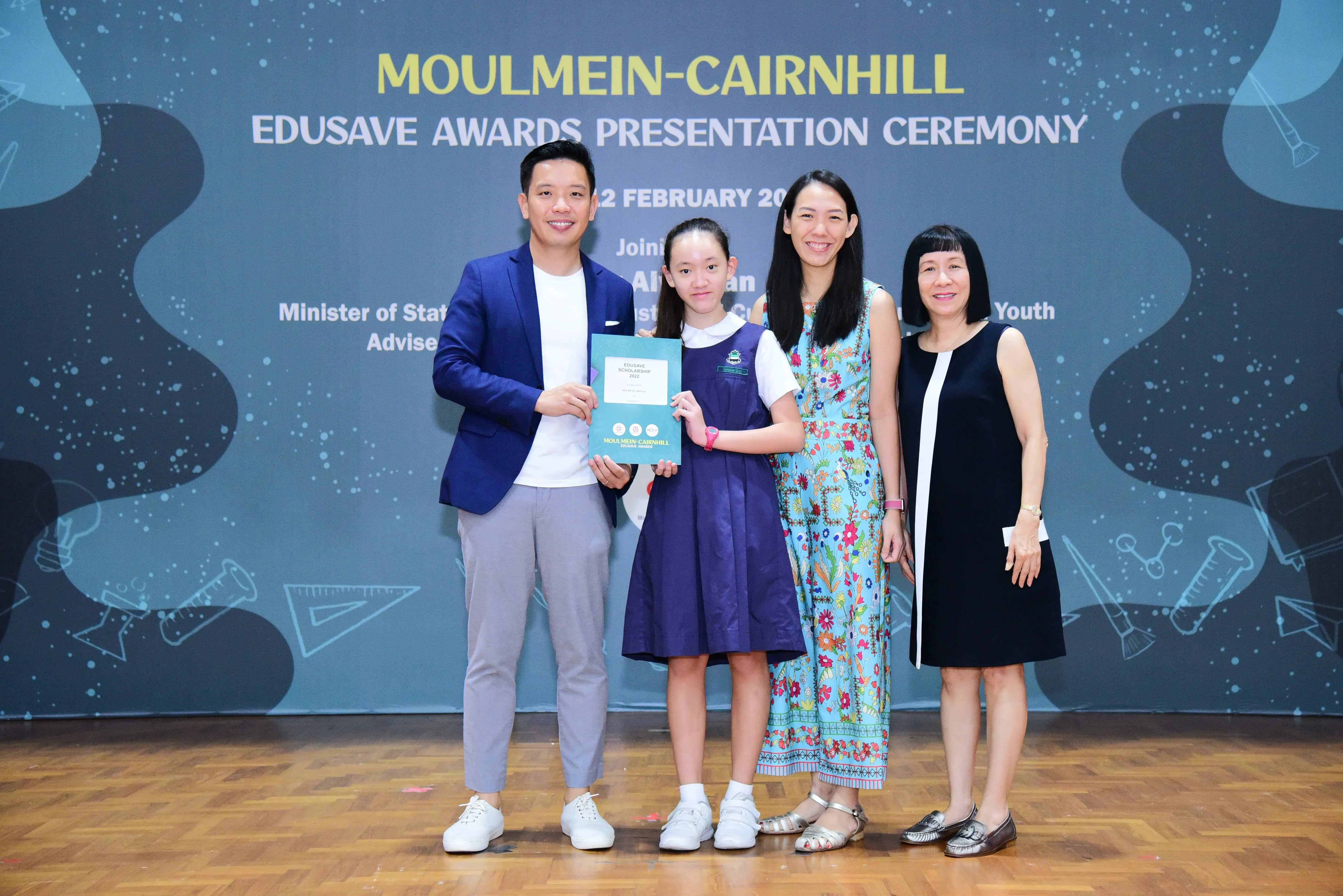Four people pose at the Moulmein-Cairnhill Edusave Awards Presentation Ceremony, with a girl holding a certificate.