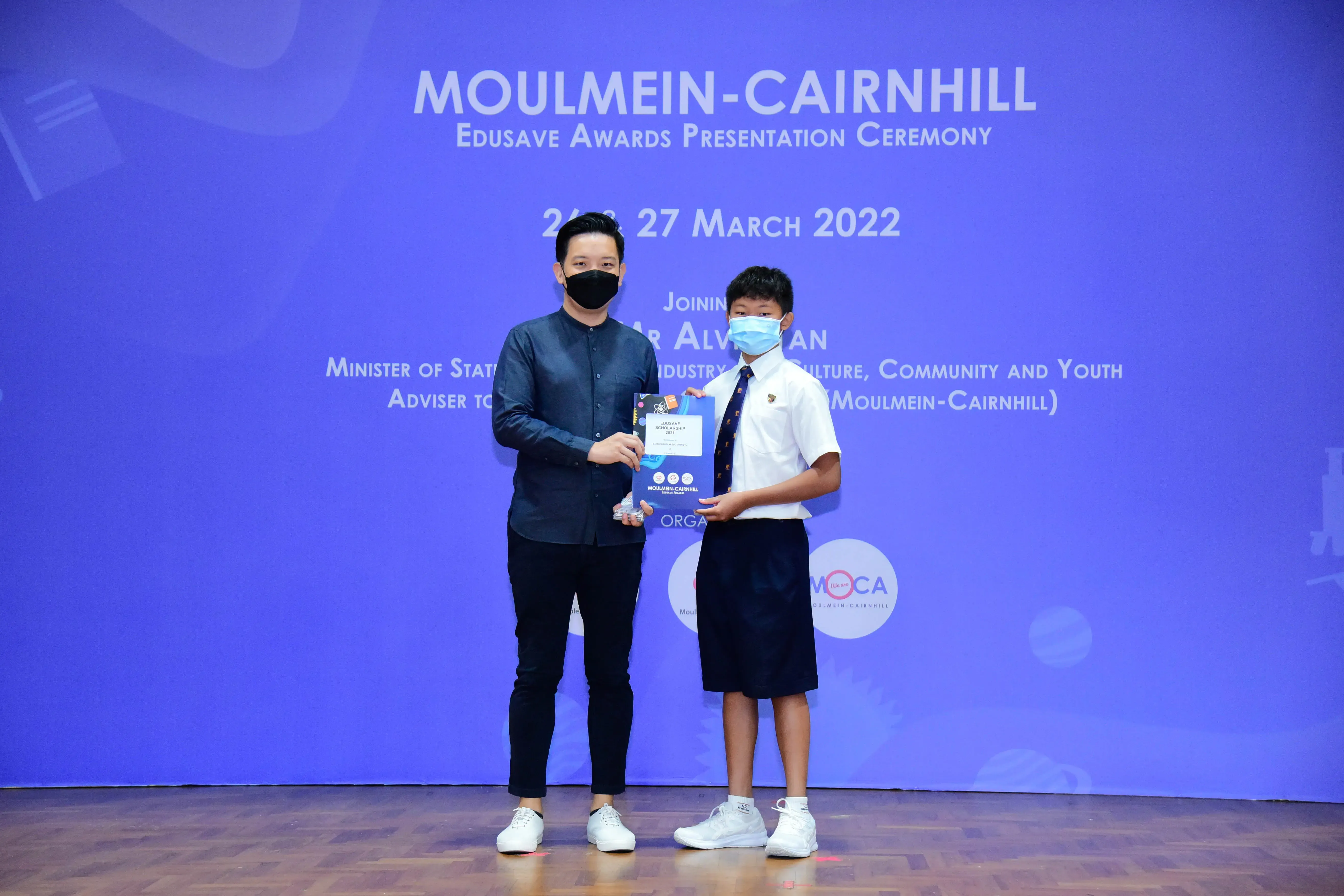 An adult and a young student in school uniform hold an award together at the Moulmein-Cairnhill Edusave Awards Ceremony 2022.