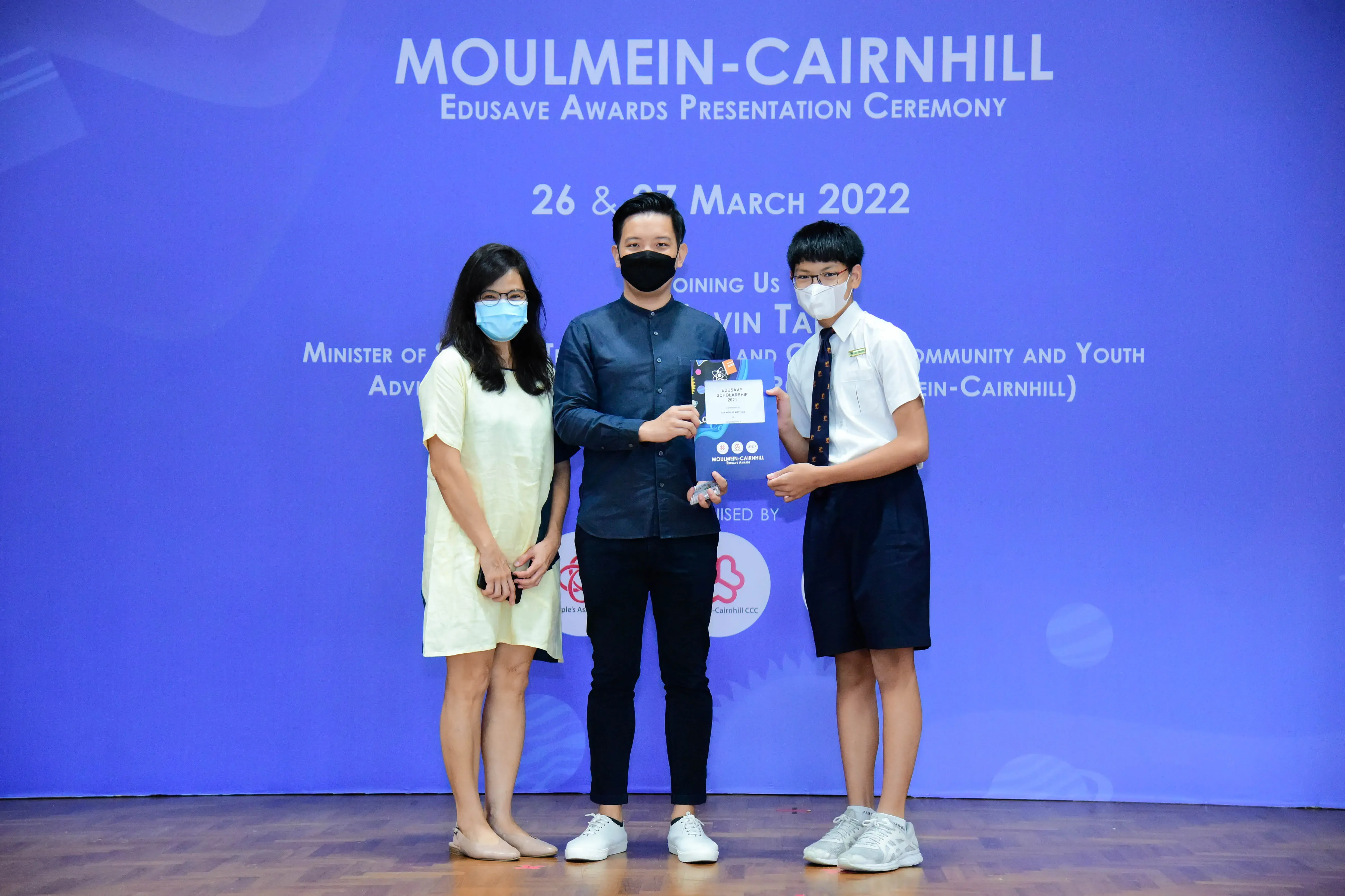 Three masked people pose on a stage at the Moulmein-Cairnhill Edusave Awards Ceremony, March 2022.