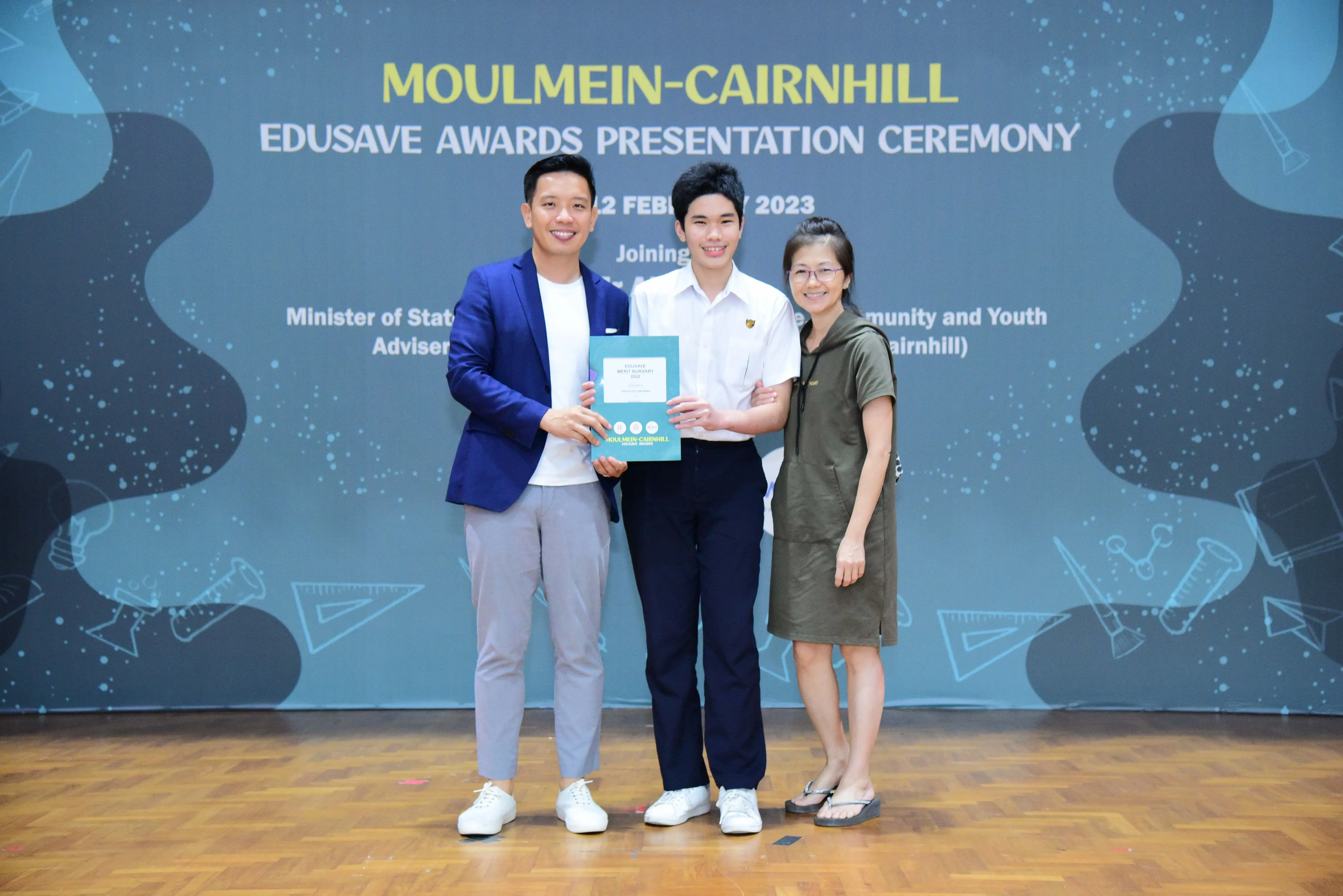 Three people smile on stage at the Moulmein-Cairnhill Edusave Awards Presentation Ceremony, holding a certificate.