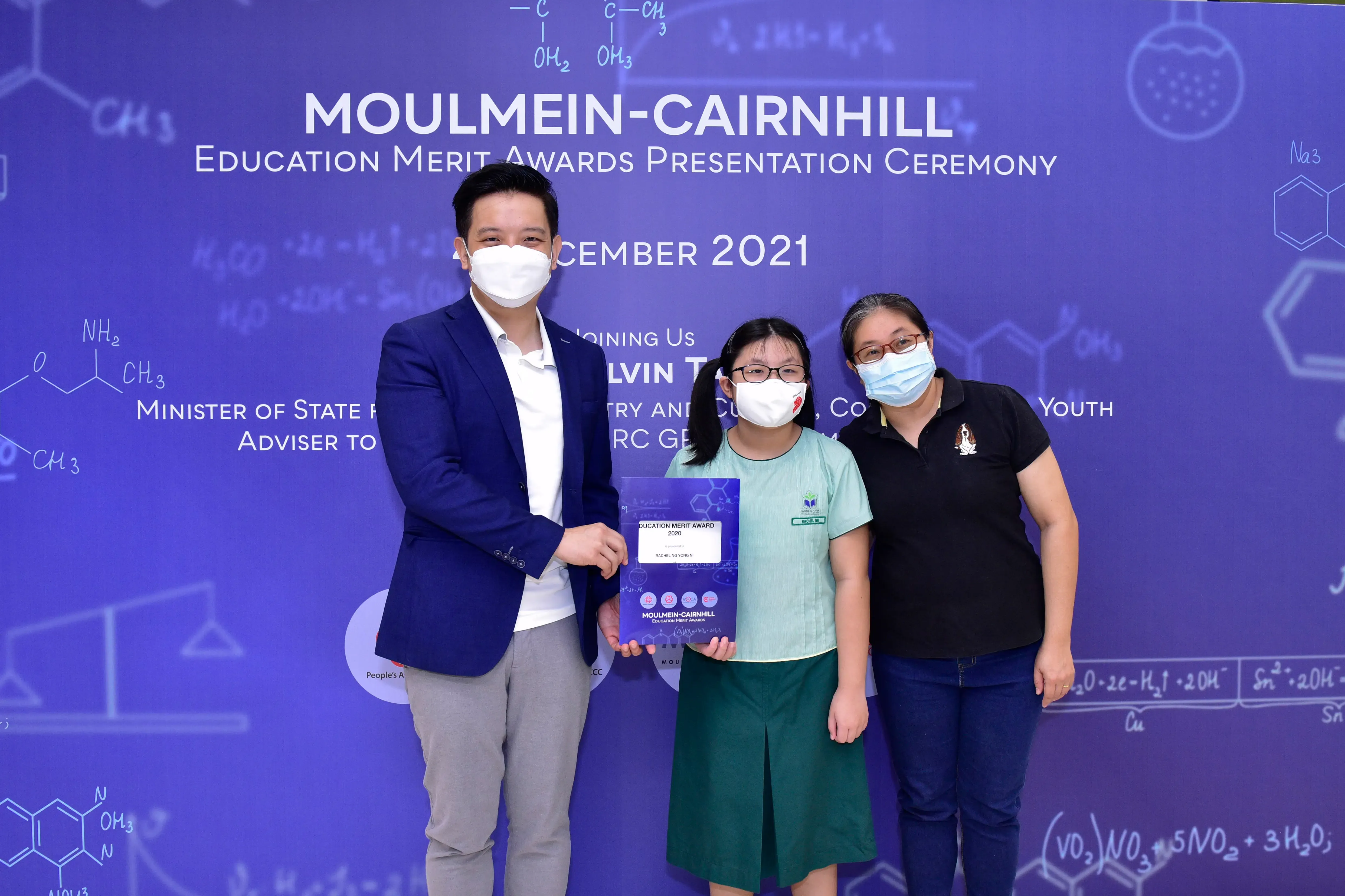 Three masked people pose at the Moulmein-Cairnhill Education Merit Awards Presentation Ceremony 2021, holding a certificate.