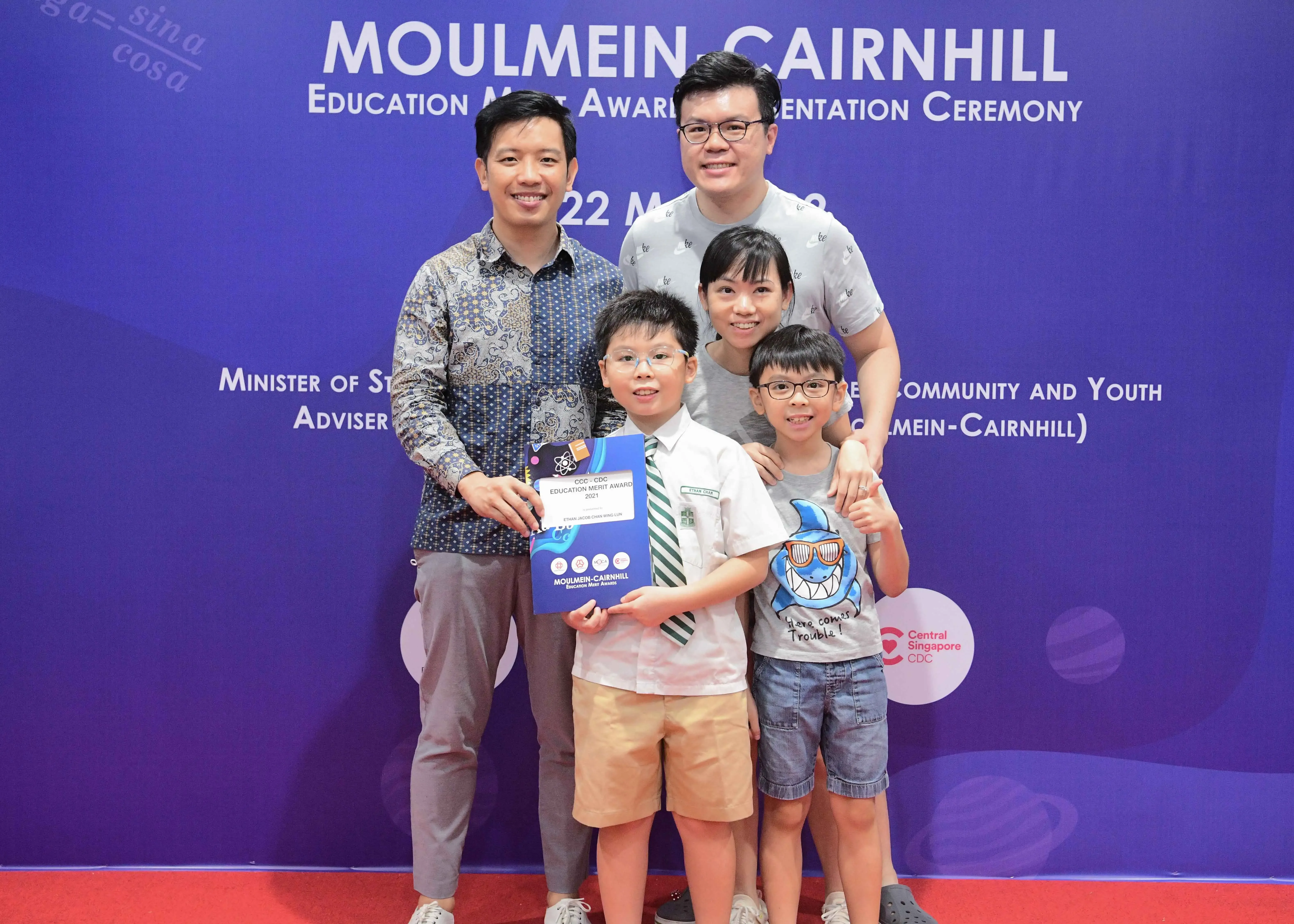 Family of four posing with an Education Merit Award certificate at the Moulmein-Cairnhill ceremony backdrop.