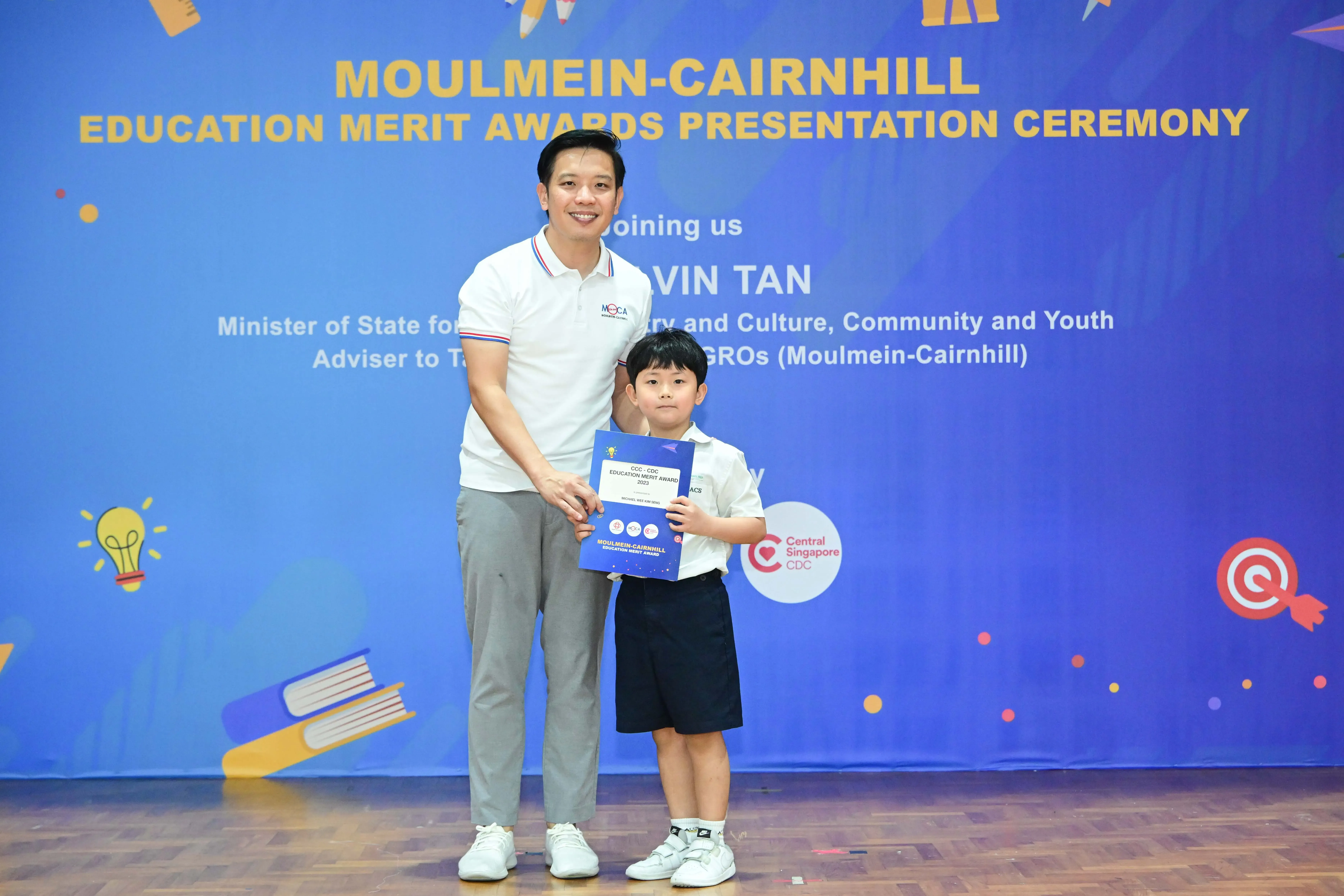 Man and young boy pose together holding a blue certificate at Moulmein-Cairnhill Education Merit Awards ceremony.