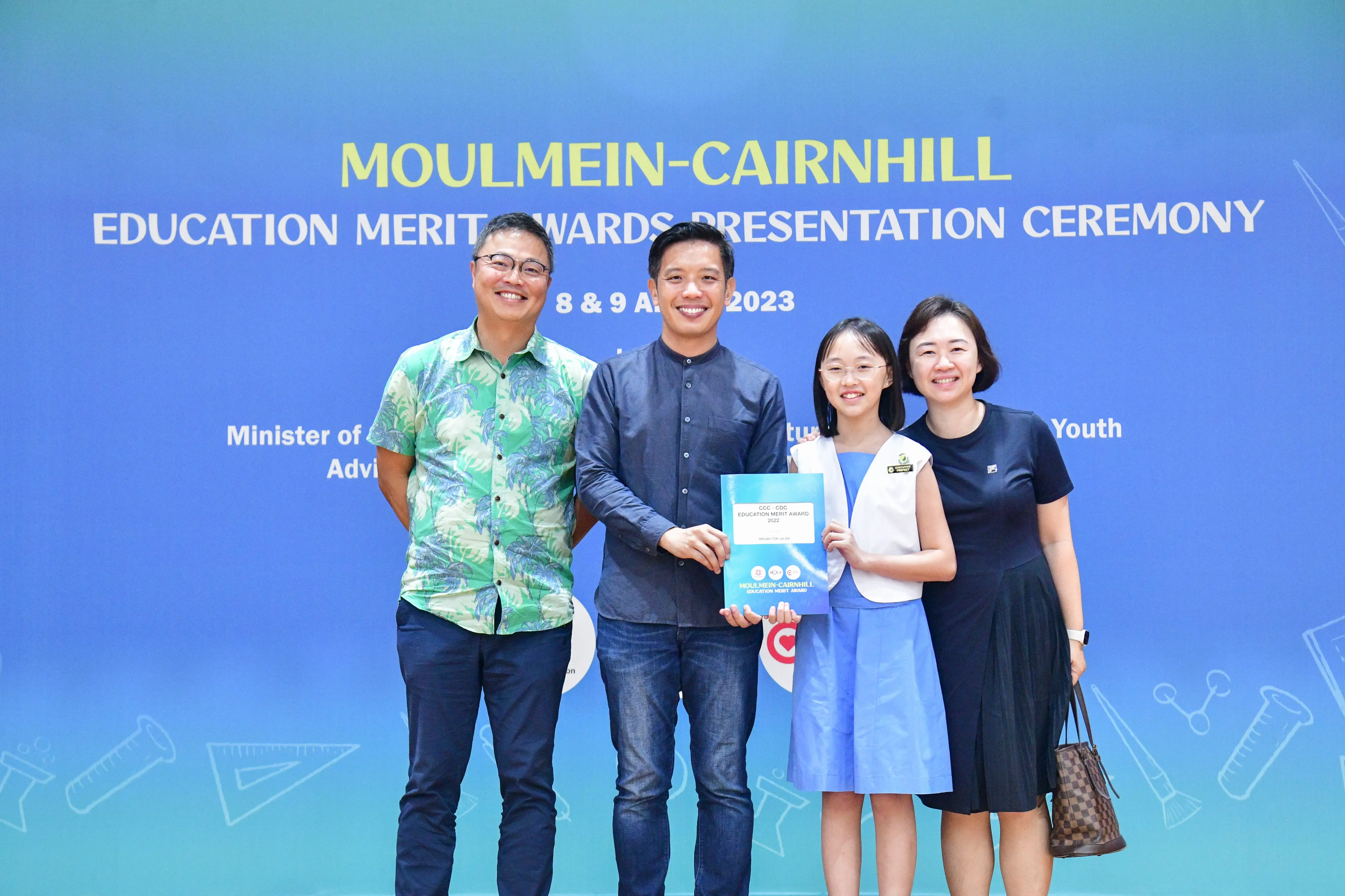 Four people pose in front of a Moulmein-Cairnhill Education Merit Awards Presentation Ceremony banner in 2023.