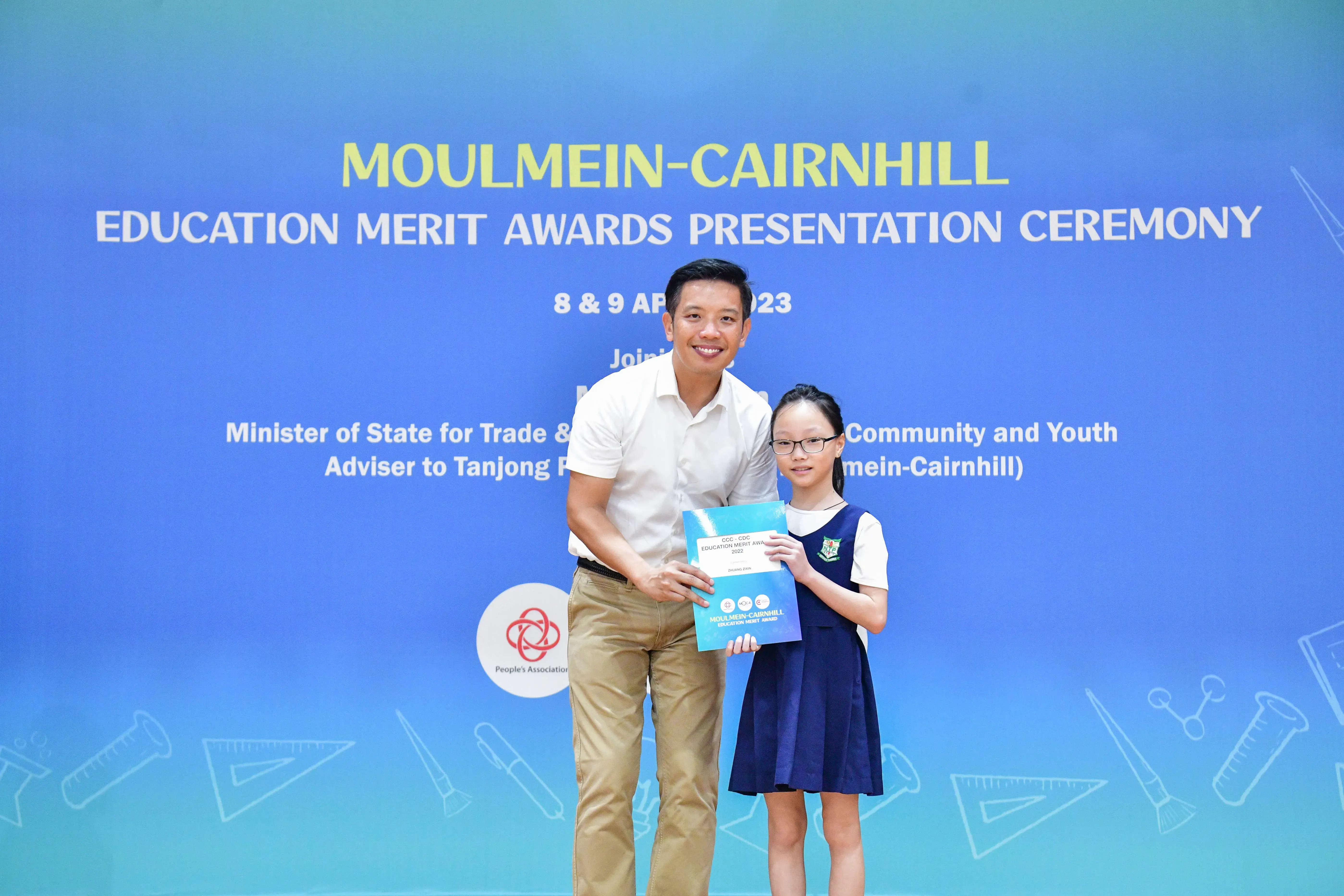 A man and a young girl holding an award certificate stand before a Moulmein-Cairnhill Education Merit Awards banner.