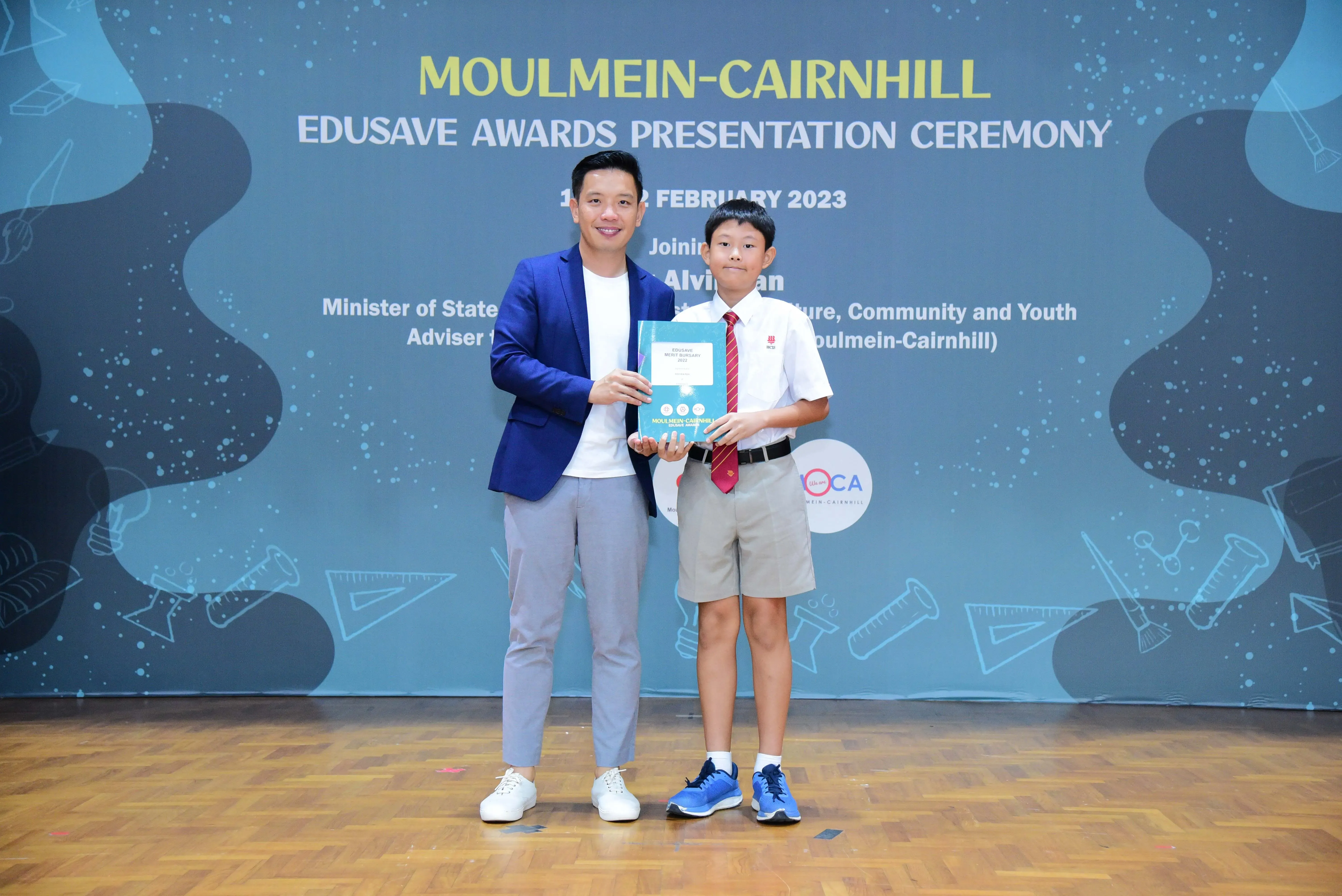 An adult in a blue blazer and a young student in uniform hold a book at the Moulmein-Cairnhill Edusave Awards 2023.