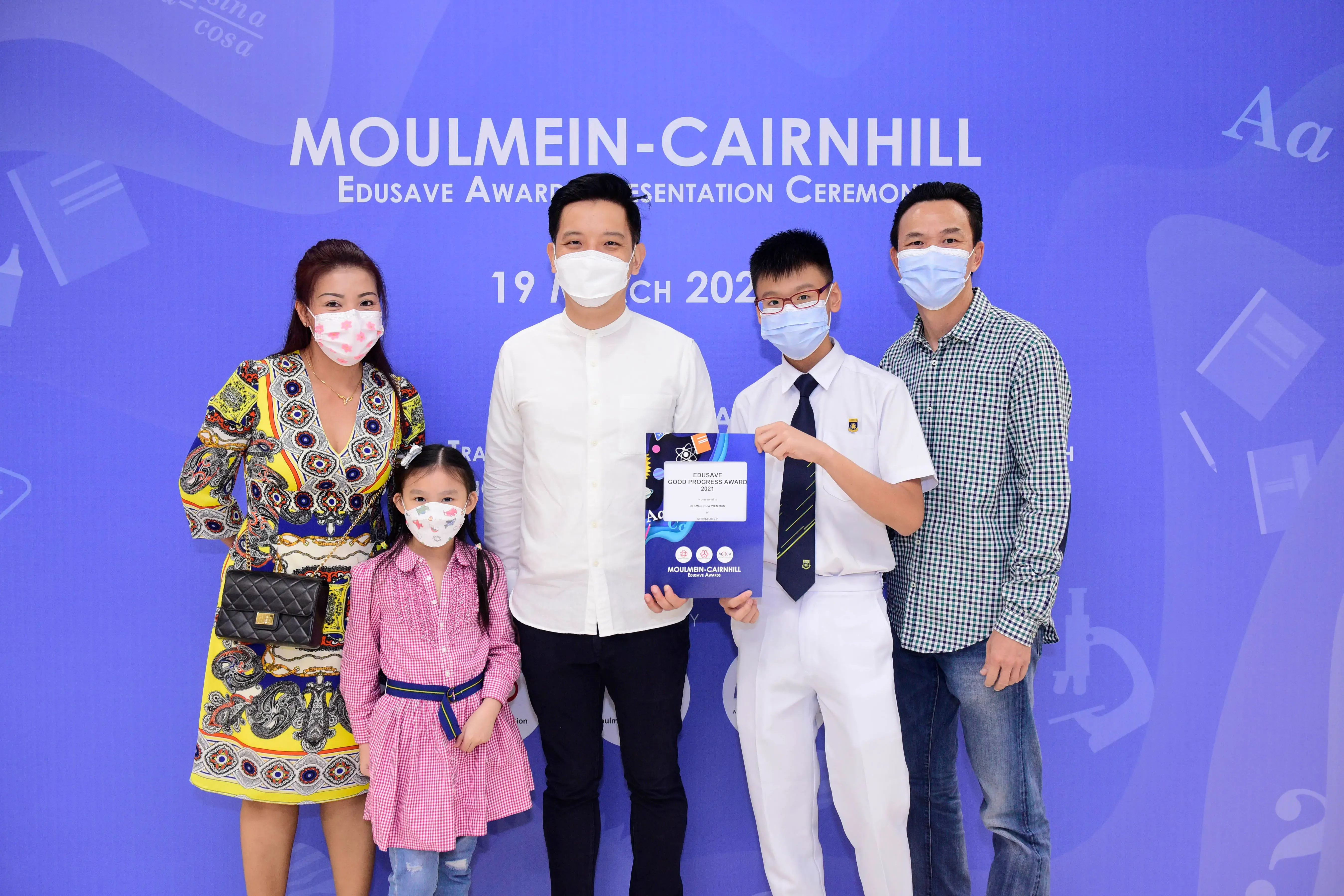 Five masked people pose at the Moulmein-Cairnhill Edusave Awards Ceremony, with a boy holding an award certificate.