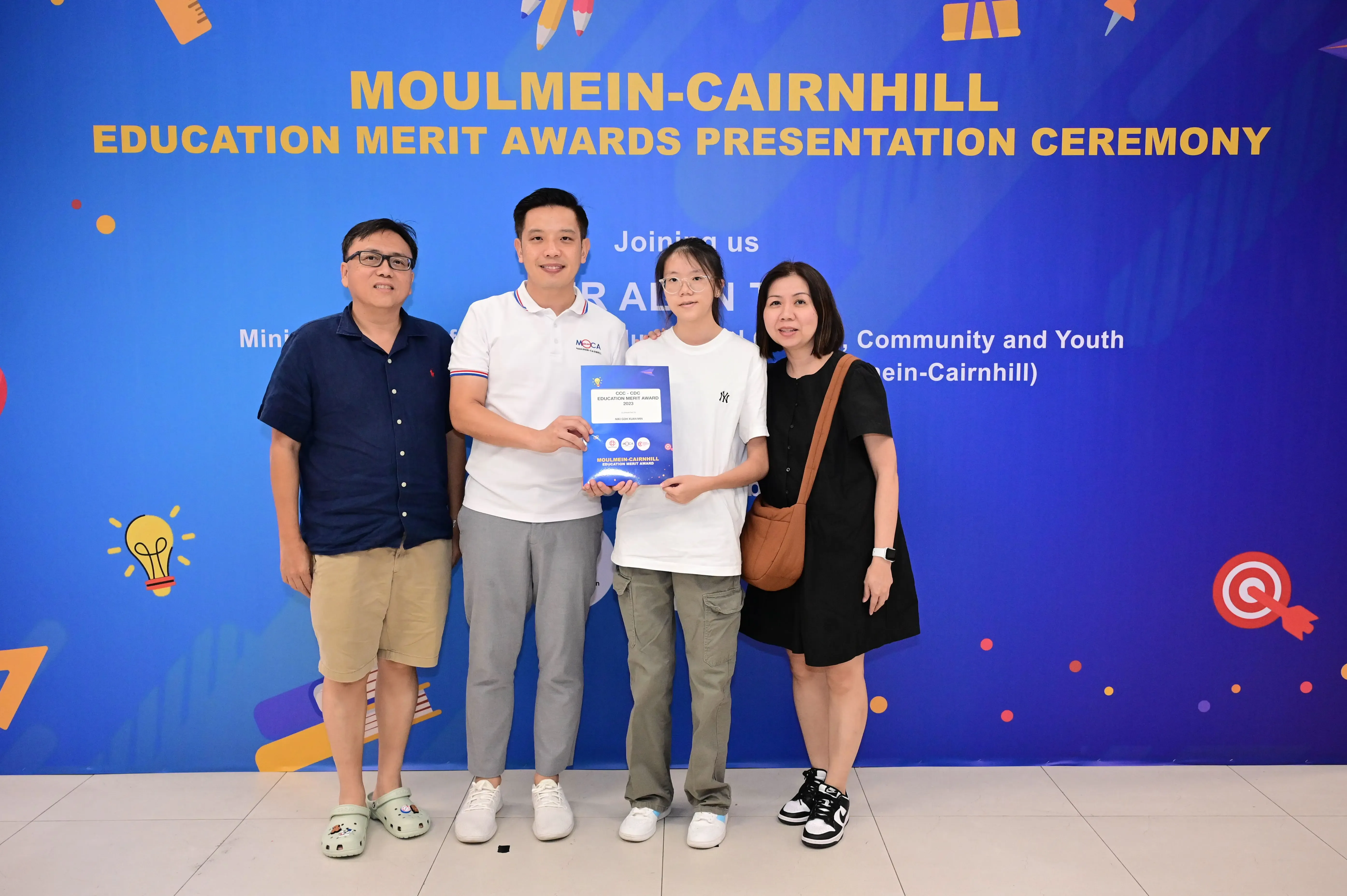 Four people pose holding a certificate at the Moulmein-Cairnhill Education Merit Awards Presentation Ceremony backdrop.