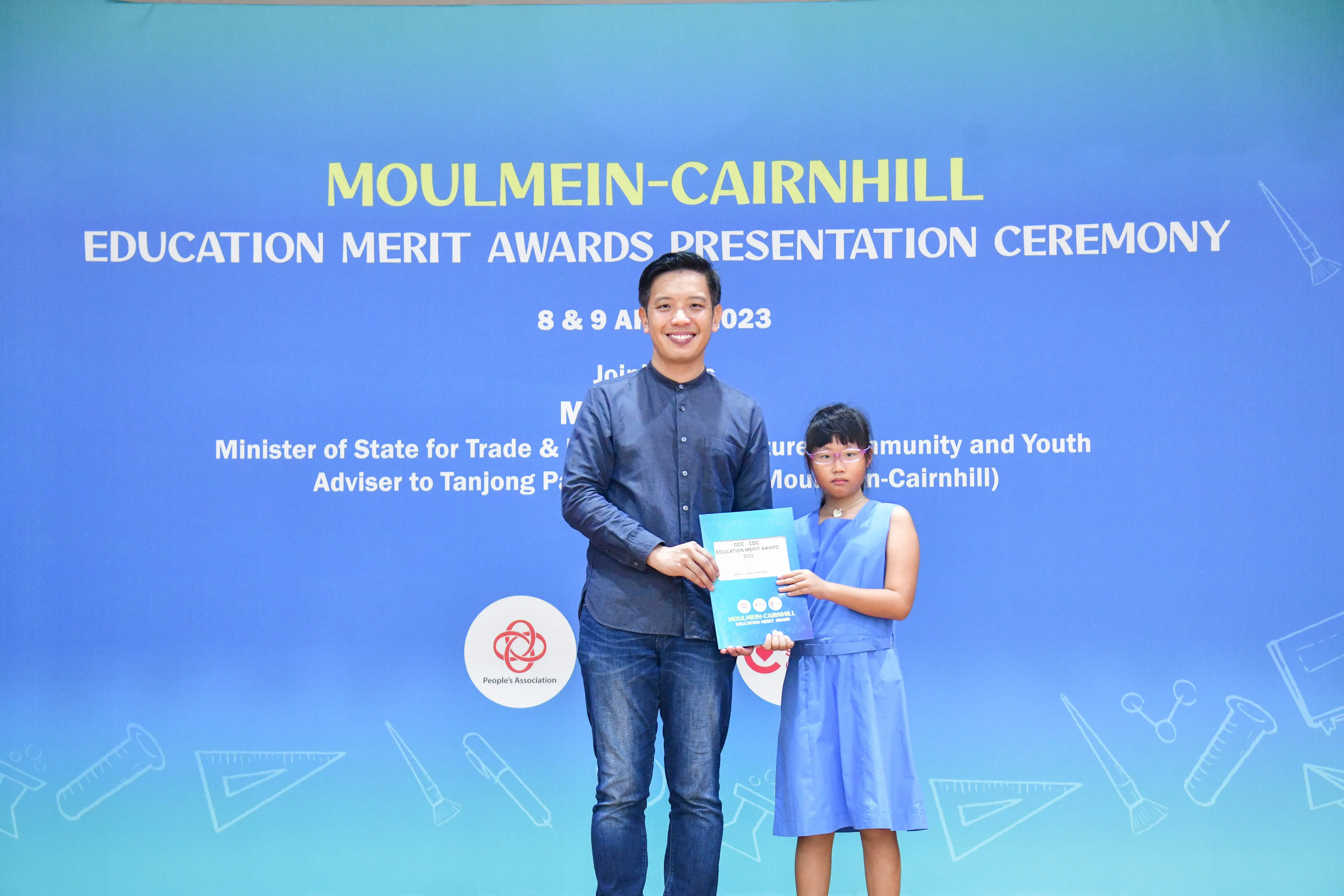A man and a young girl hold a certificate together in front of a Moulmein-Cairnhill Education Merit Awards banner.