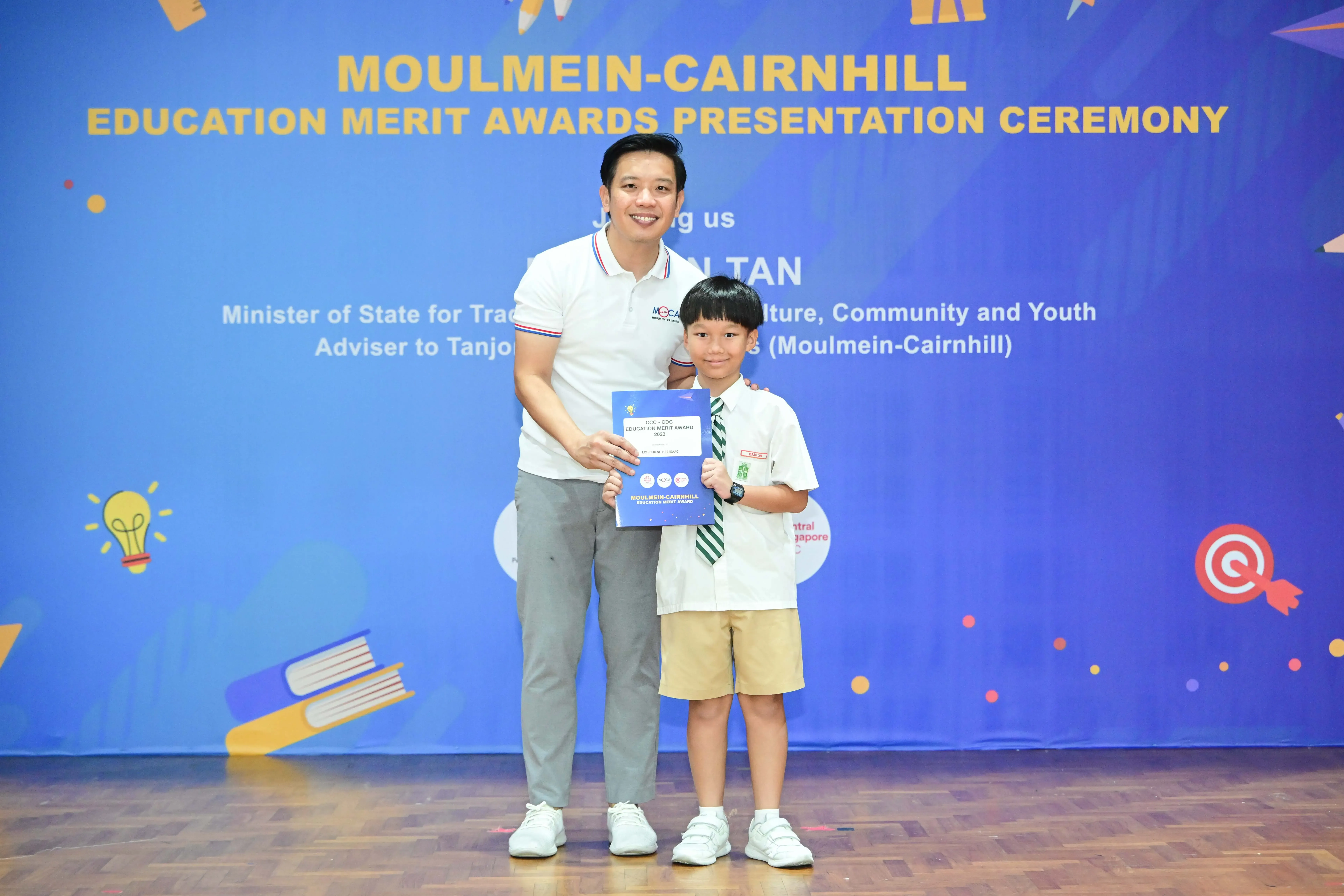 An adult and a young boy in school uniform hold a certificate at the Moulmein-Cairnhill Education Merit Awards ceremony.