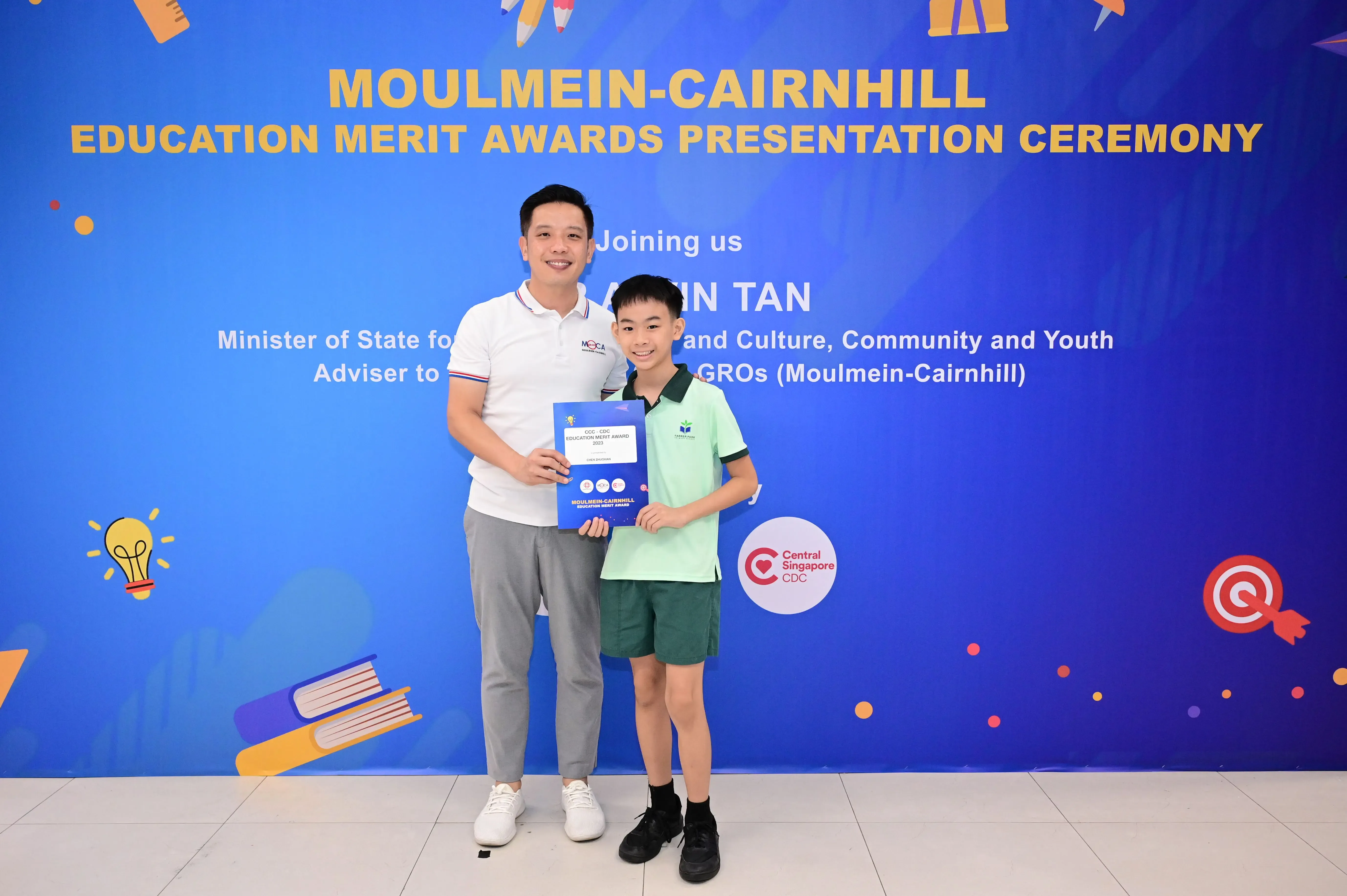 A man and a boy pose holding an award certificate in front of a Moulmein-Cairnhill Education Merit Awards banner.