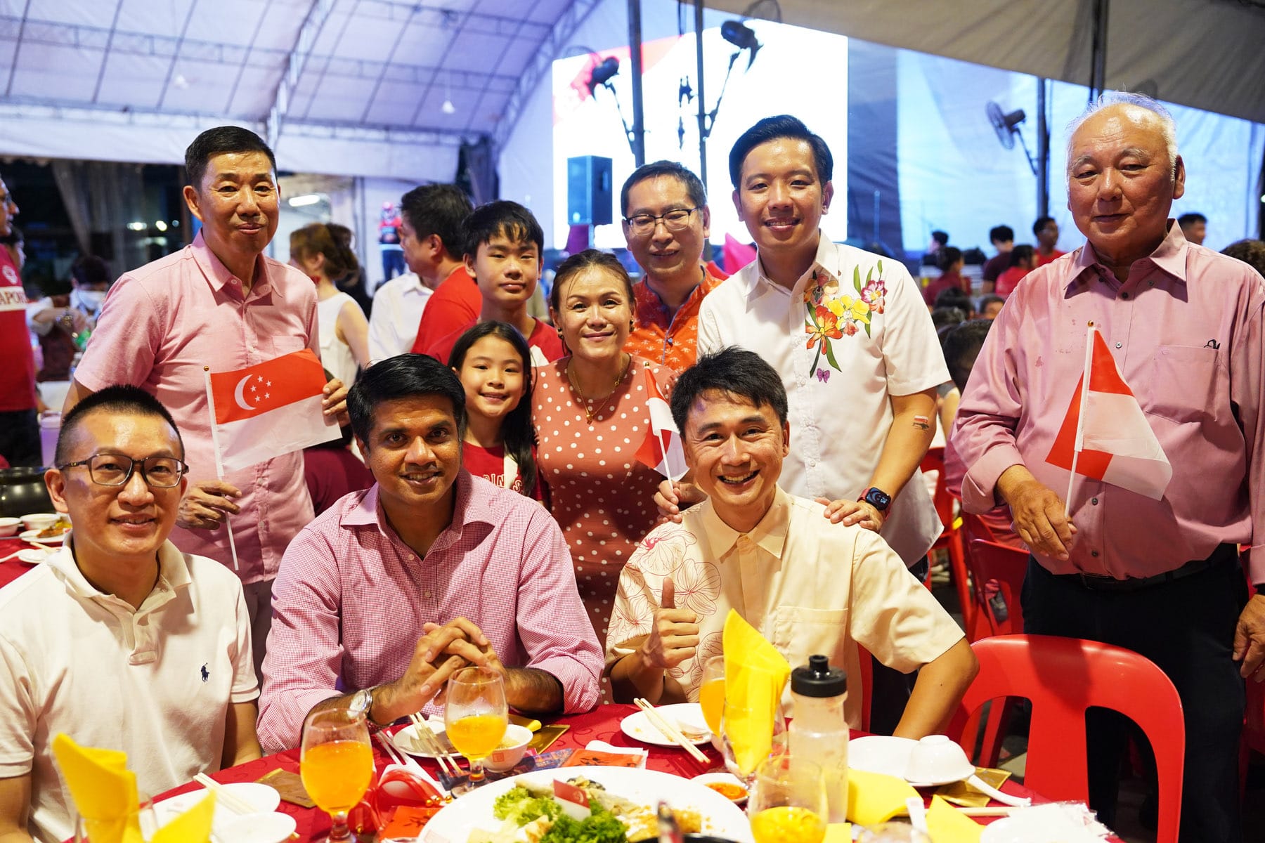 A group of smiling people pose at a festive dinner table, holding small Singapore flags, wearing red and pink outfits.