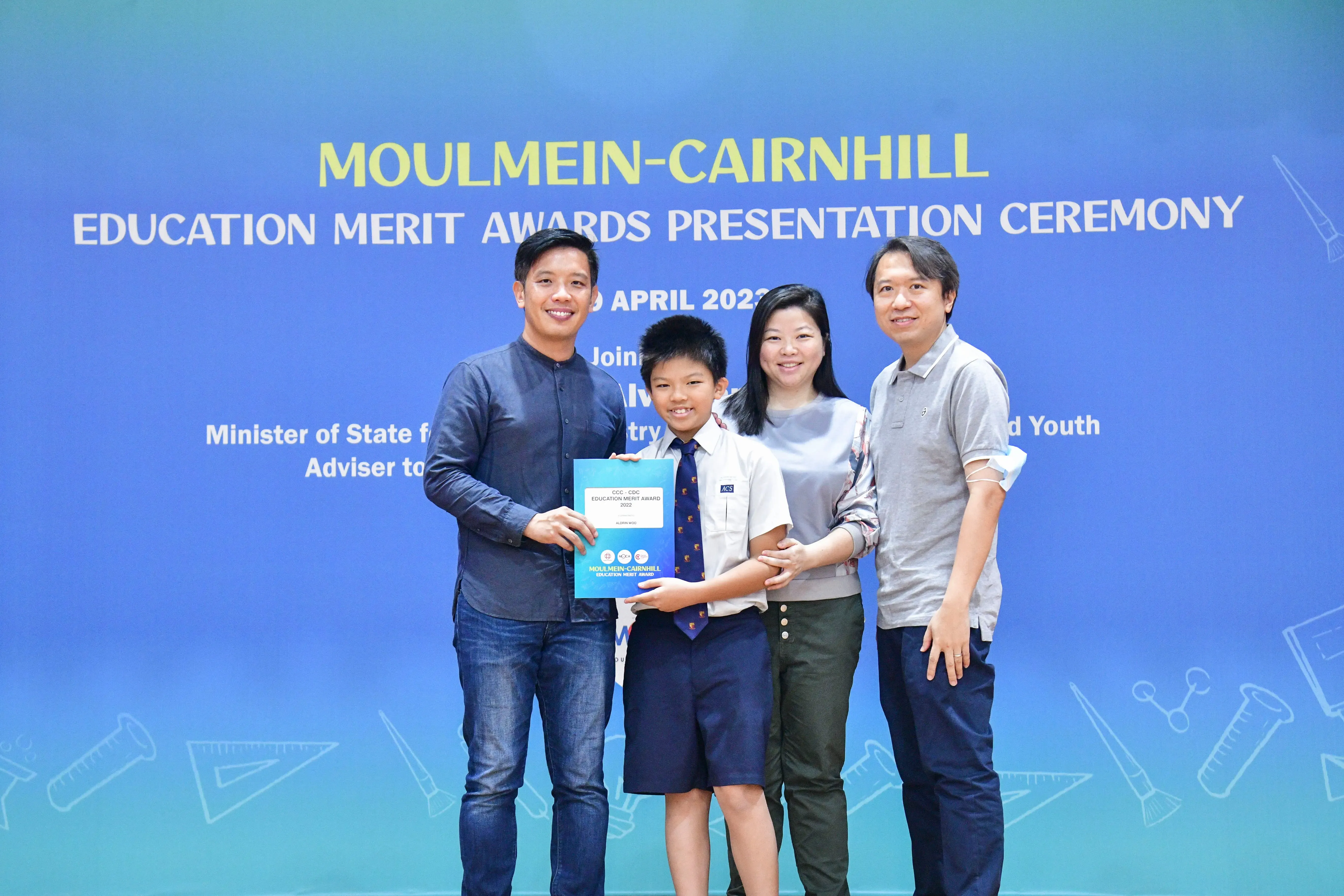 Four people pose at the Moulmein-Cairnhill Education Merit Awards Presentation Ceremony, with a boy holding an award certificate.