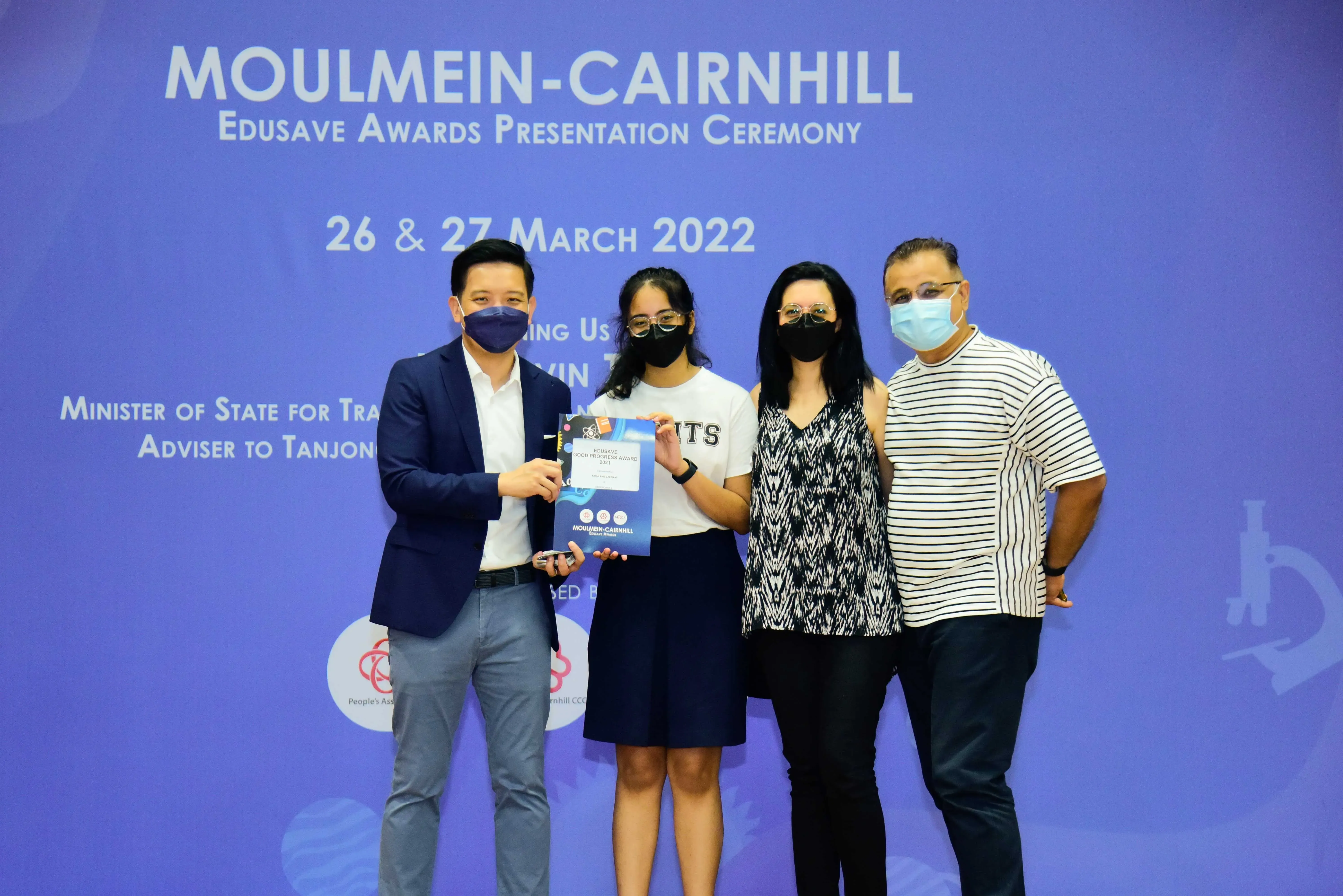 Four masked people pose at the Moulmein-Cairnhill Edusave Awards Ceremony, with a girl holding an award certificate.