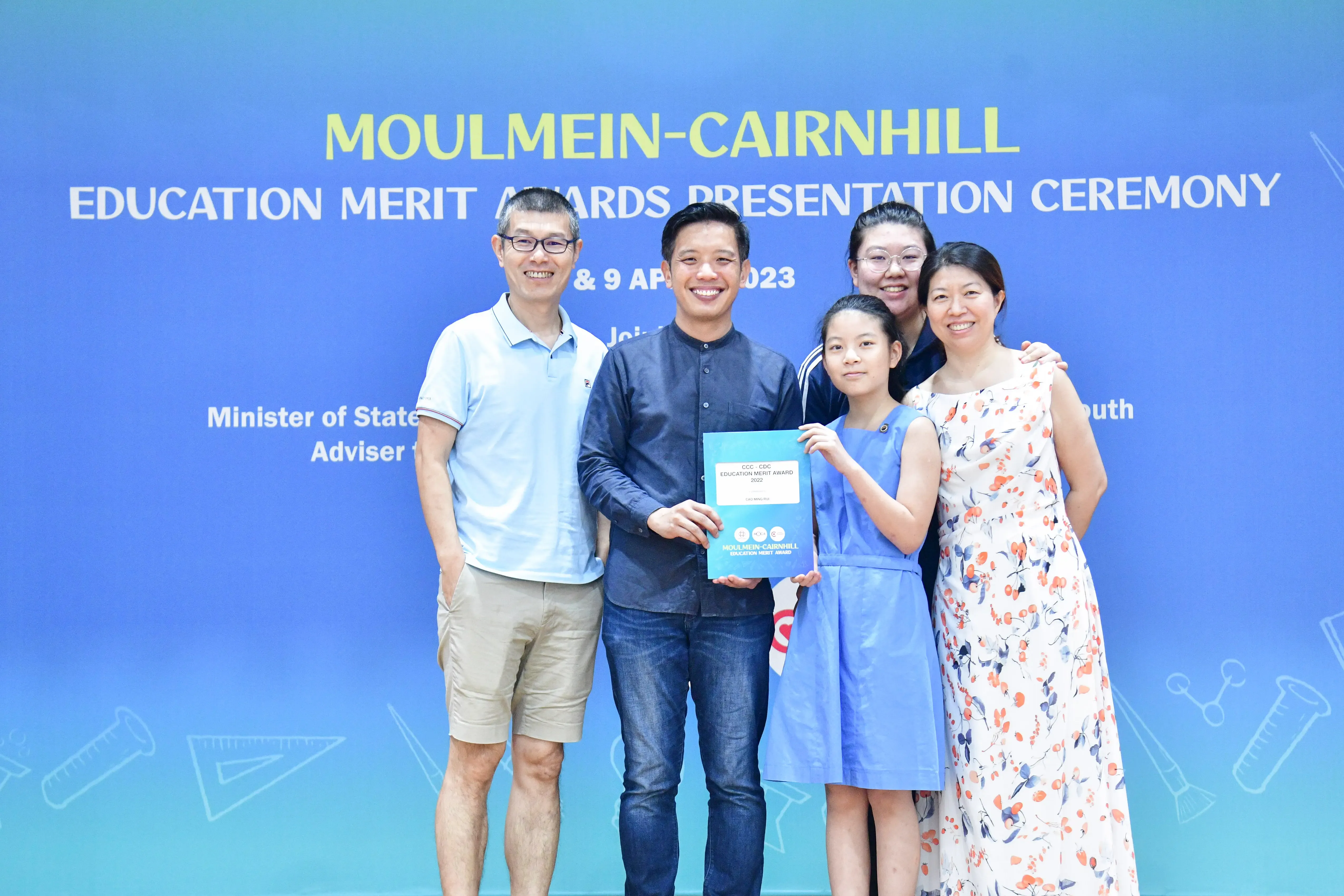 Five people pose smiling at the Moulmein-Cairnhill Education Merit Awards Presentation Ceremony, holding a teal award certificate.